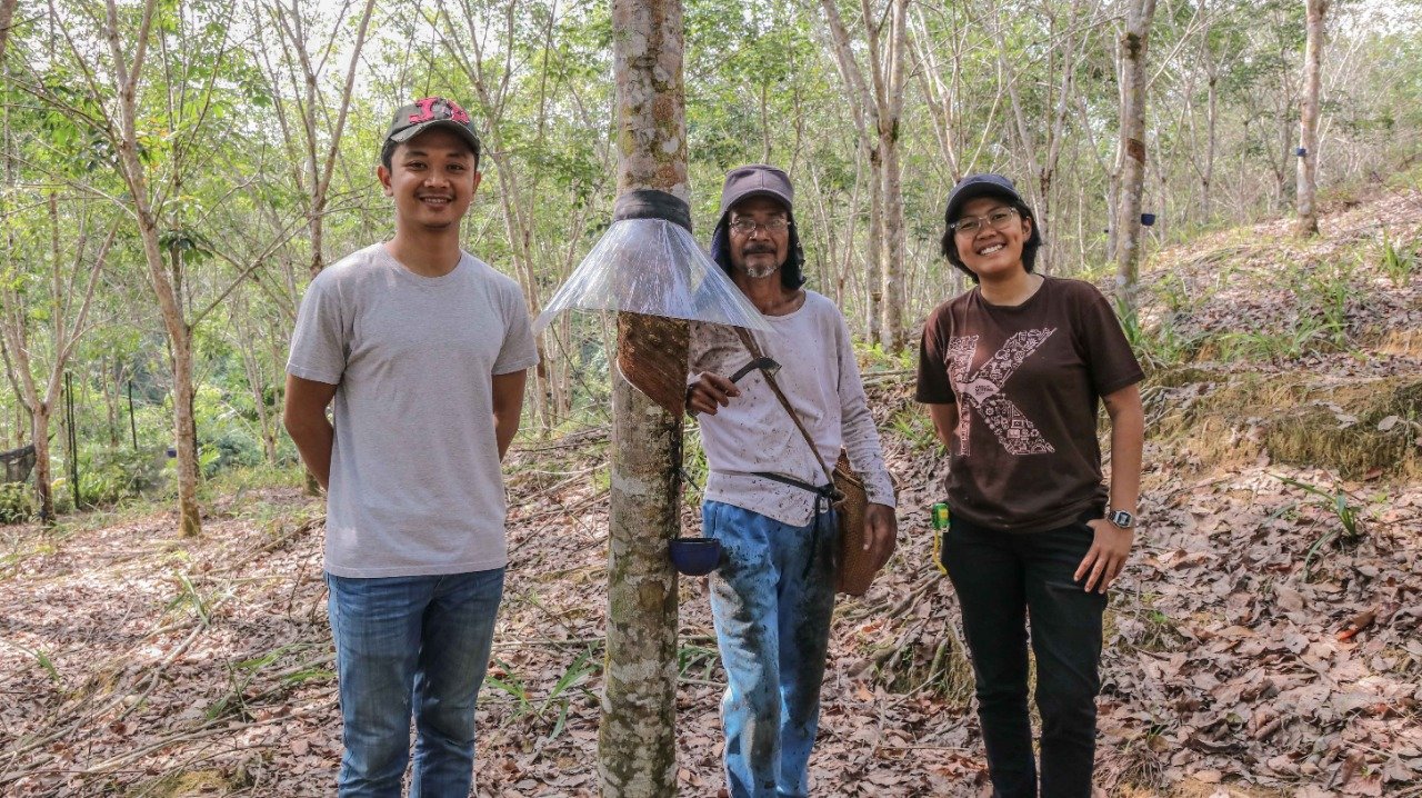 Three people standing in a forest, smiling at the camera. The person in the middle is holding a tool and has a large plastic umbrella over a tree branch.