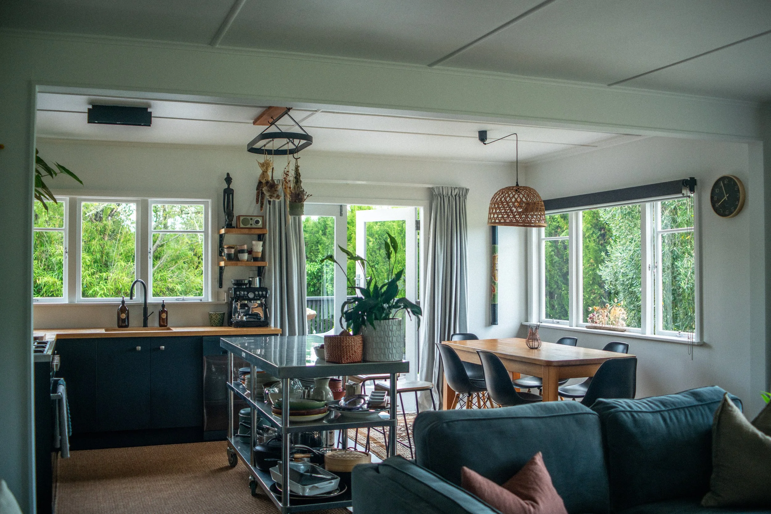 Living room and kitchen area with large windows, dining table, sofa, plant, and various decor items, overlooking a green outdoor view.