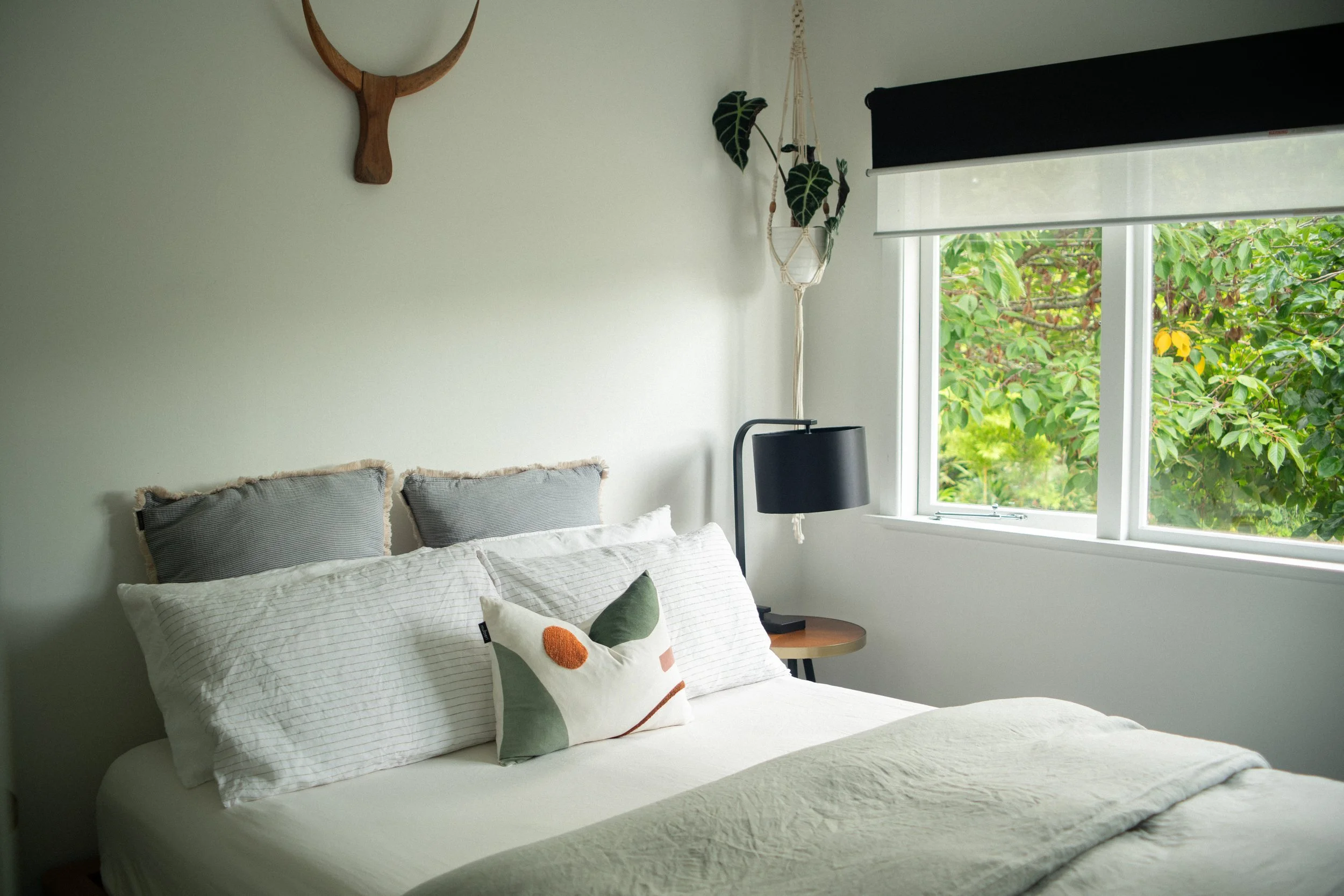 Cozy bedroom with white bed, gray pillows, decorative cushion, wooden side table, black lampshade, large window with greenery outside, hanging plant, wall decoration resembling tree branches.