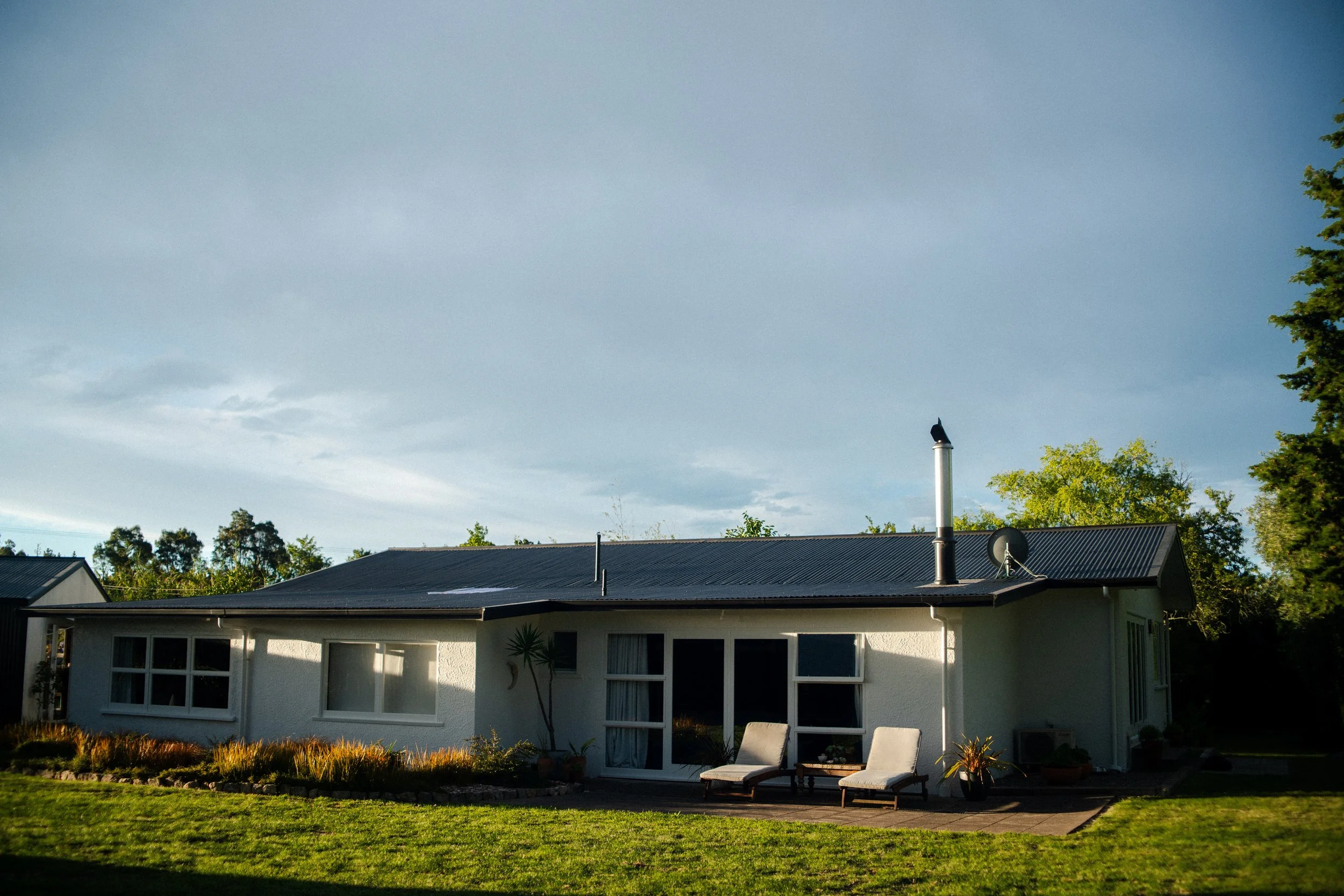 A single-story house with a black metal roof, white exterior walls, and multiple windows, surrounded by a well-maintained lawn and trees under a partly cloudy sky.