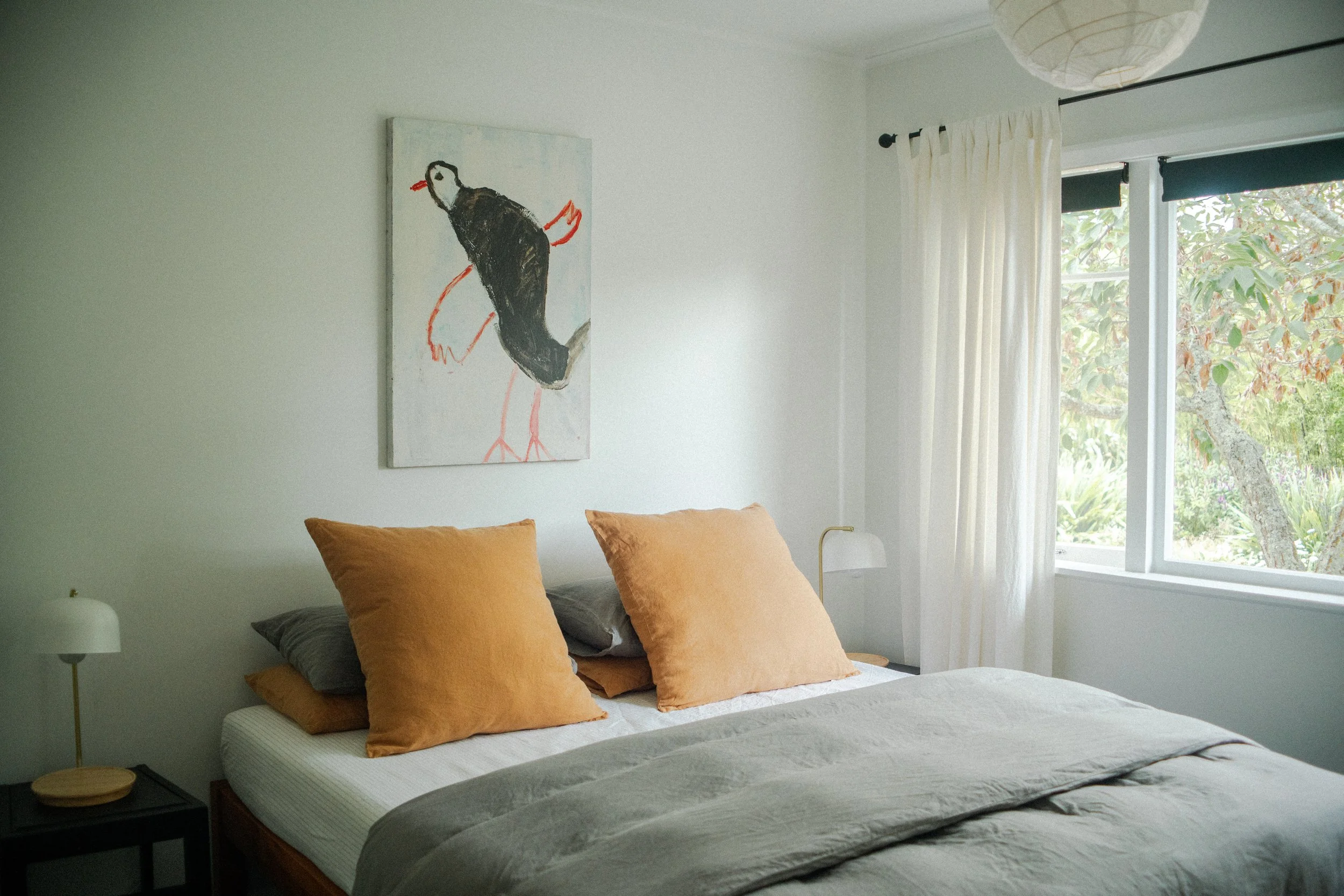 A neatly made bed with beige and gray pillows, a beige blanket, and a white bedspread in a bright bedroom. There is a framed artwork of a black bird on the wall above the bed. A window with white curtains and a view of trees outside is on the right s