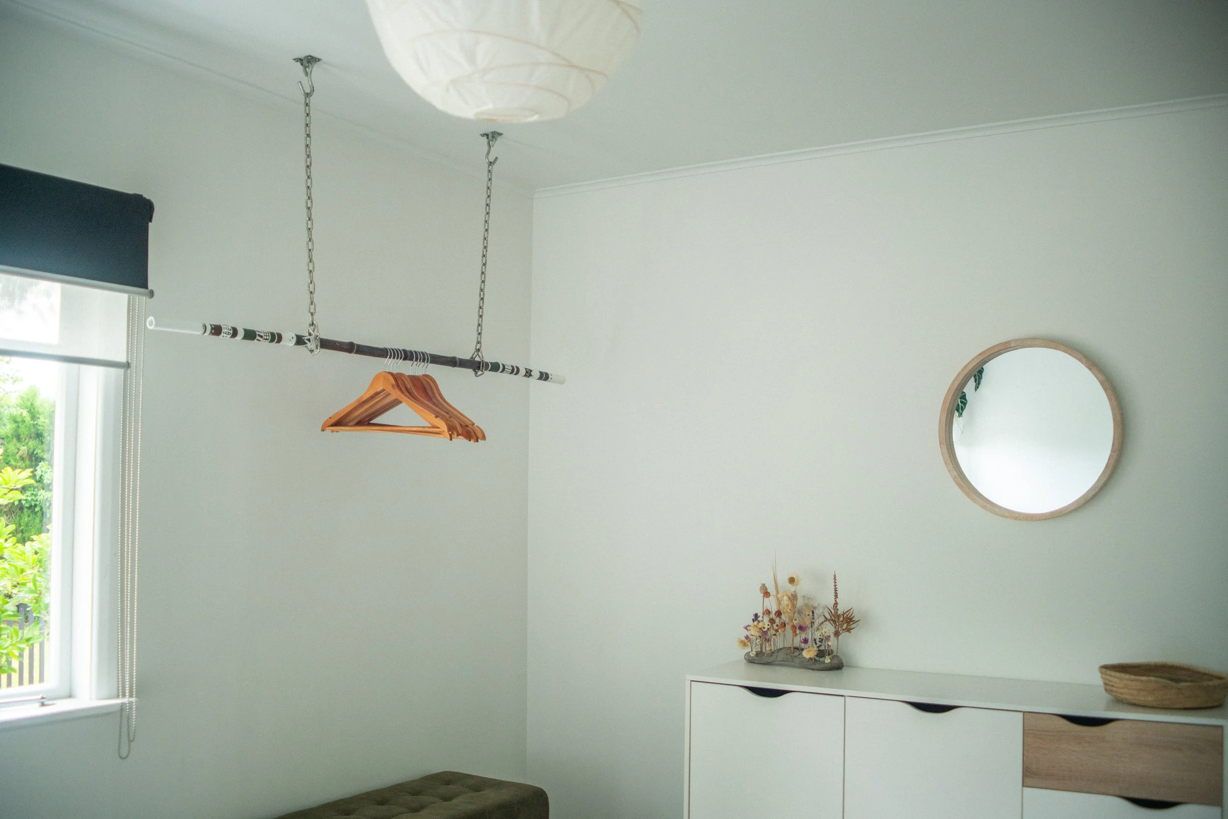A room with a window on the left, a ceiling light fixture, a hanging rod with a few wooden hangers, a mirror on the wall, a sideboard with flowers and a basket.