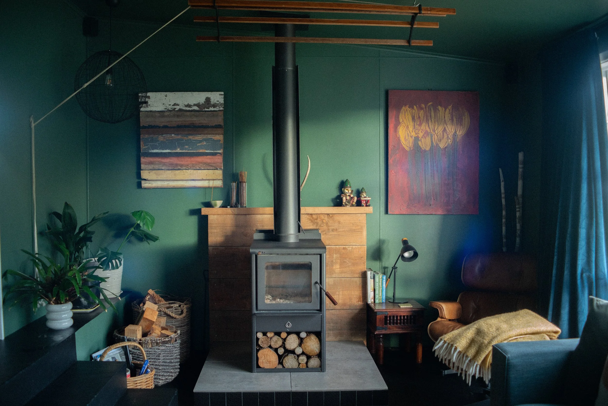 Living room with green walls, a wood stove with firewood underneath, a small side table with books and a desk lamp, an armchair with a yellow blanket, plants, colorful abstract paintings, and a window with blue curtains.