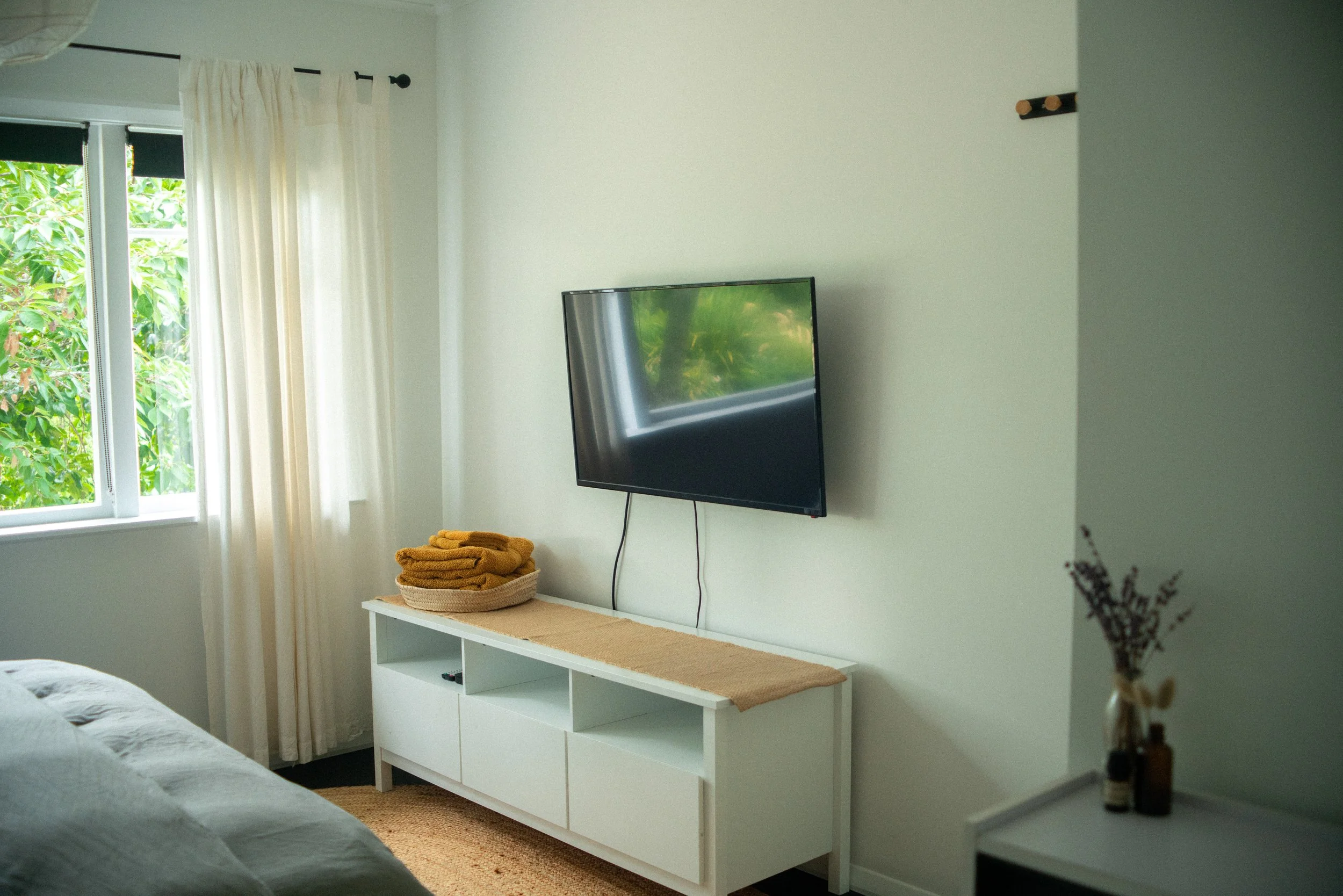 Interior of a room with a window, a mounted flat-screen TV, a white console table with folded towels, and decorative bottles with dried flowers.