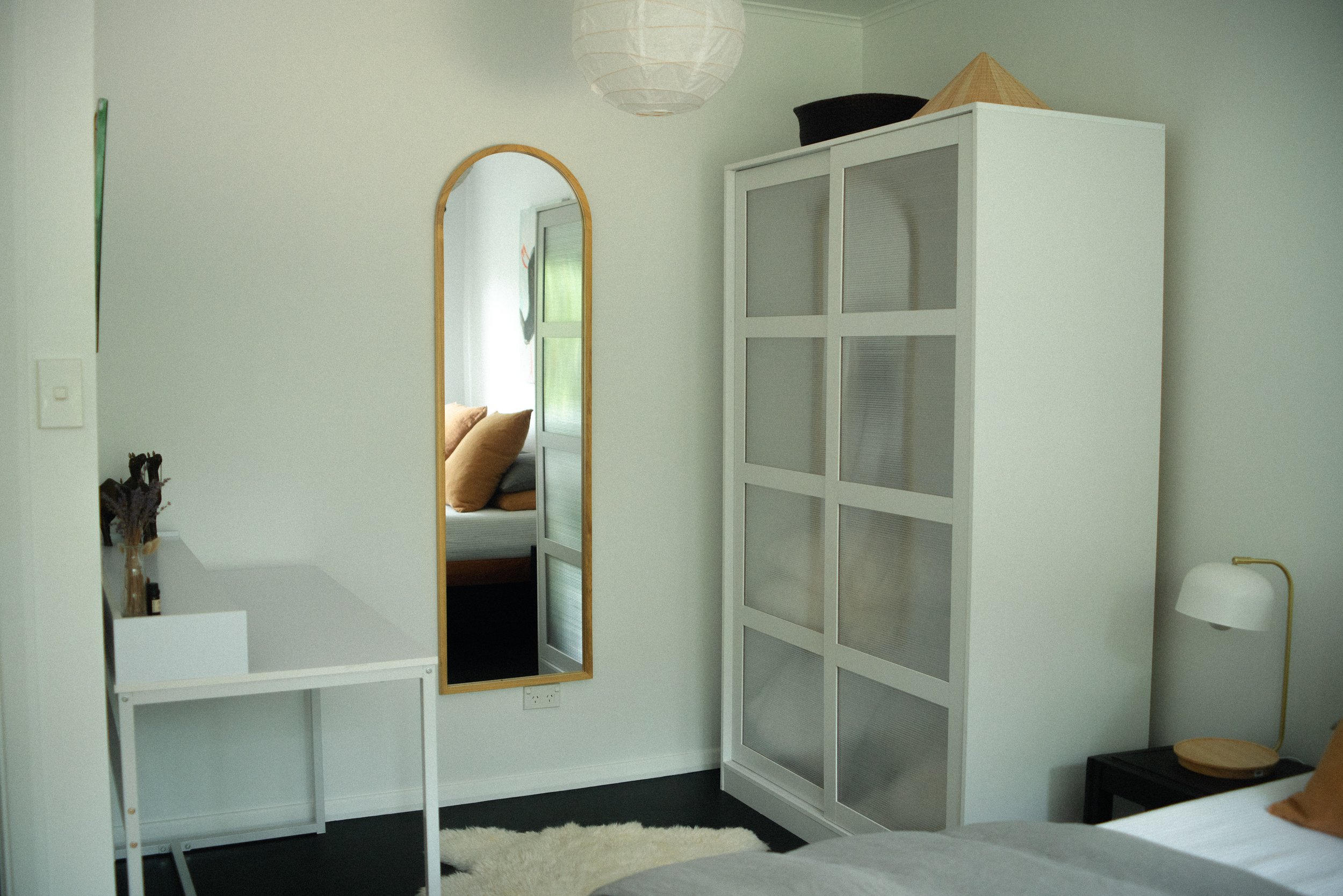 A modern bedroom with a white wardrobe, a black side table with a small lamp, a round mirror on the wall, and a bed with brown pillows.