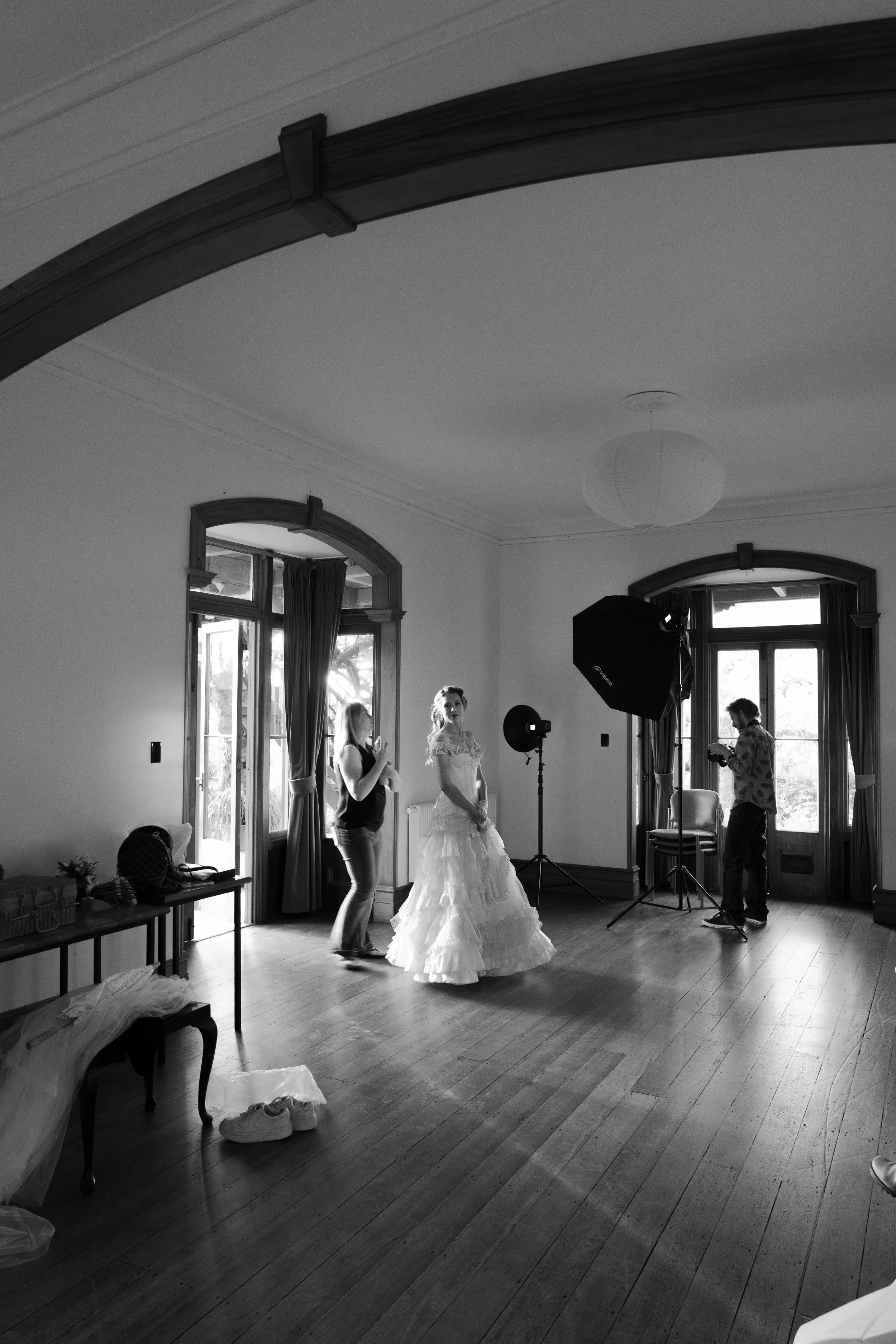 A bride in a wedding dress being prepared in a room with three windows, two are open, with curtains and a door. Two women and a man are setting up photography equipment, with one woman on her phone, another woman standing beside the bride, and the ma