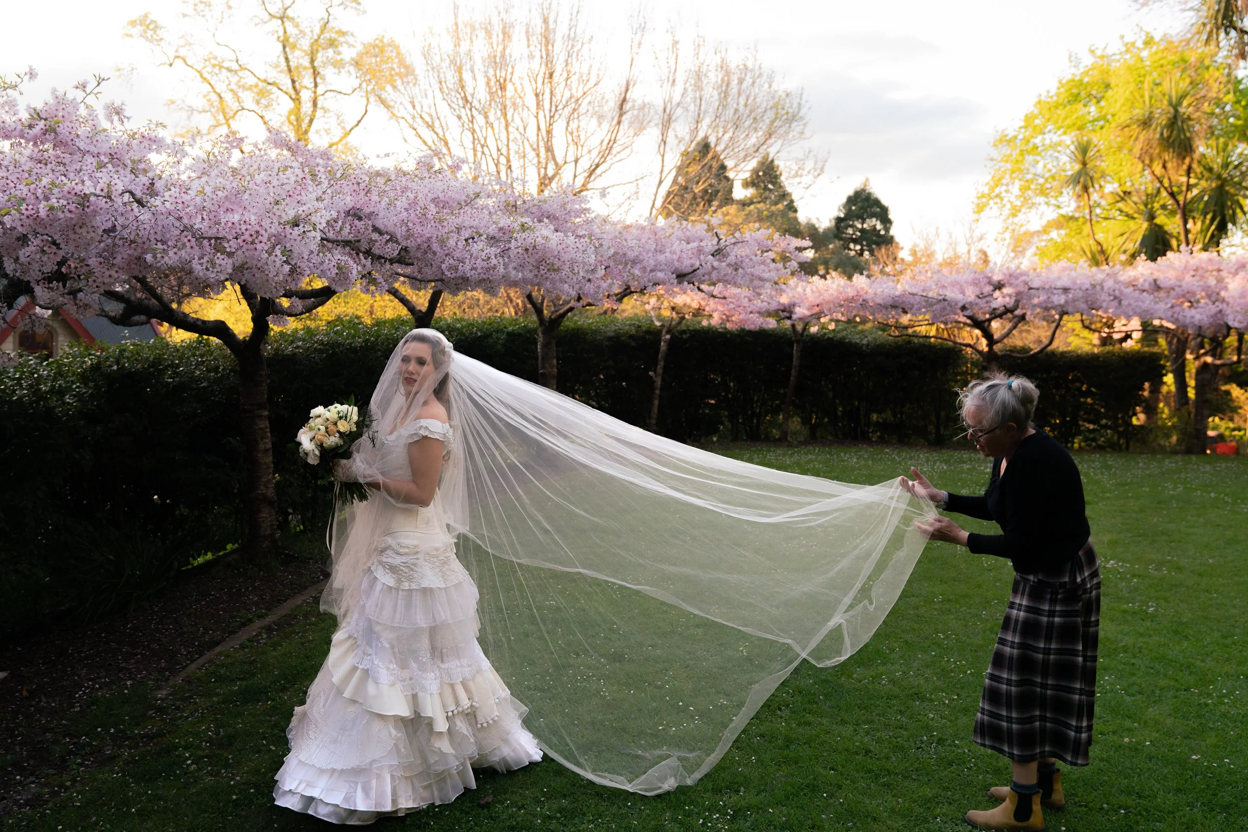 A bride in a white wedding dress holds a bouquet of flowers while an older woman helps her with her long veil in a garden with blooming cherry blossom trees.