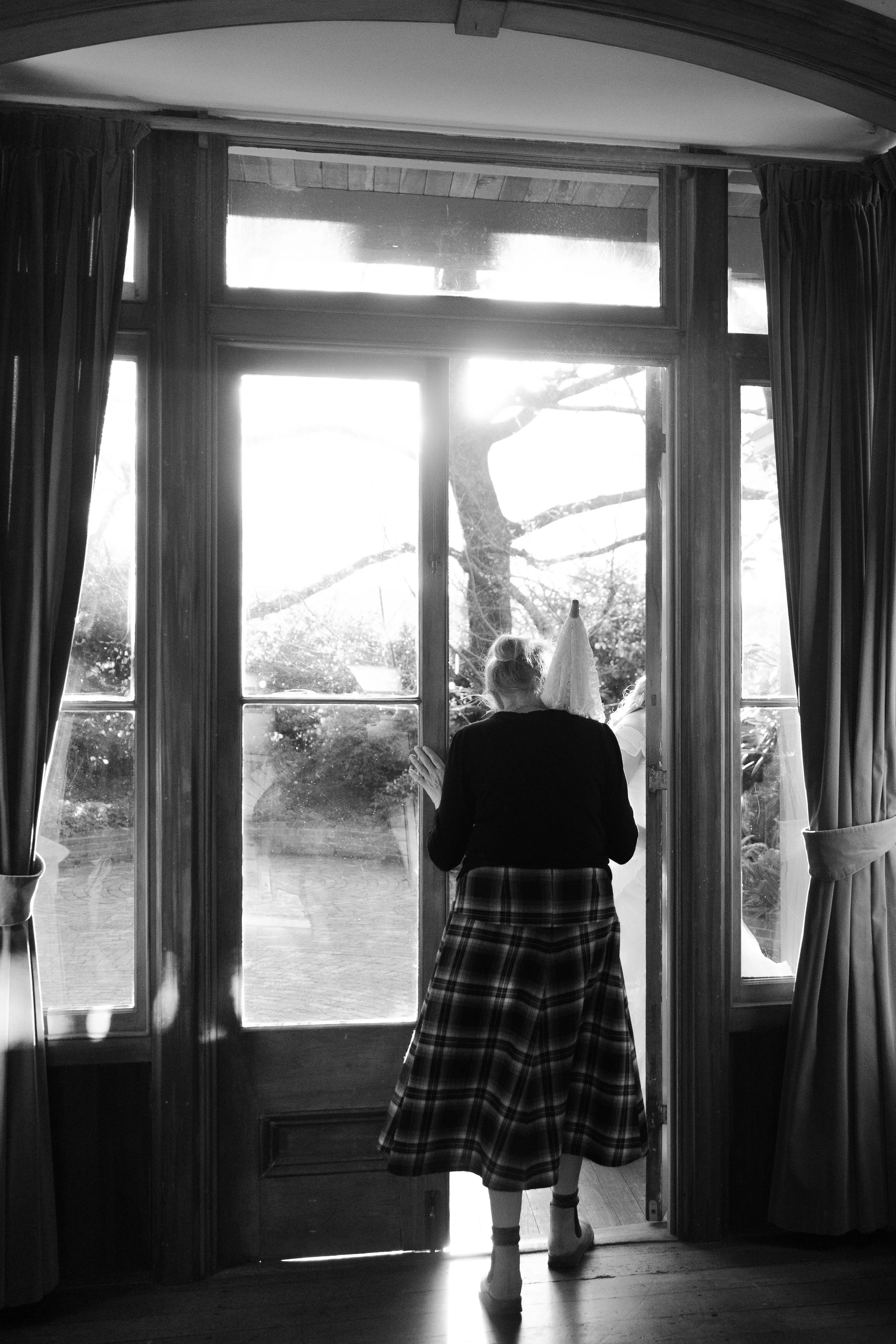 A woman wearing a plaid skirt and heels stands inside a house, looking out through a glass door at a tree outside. The scene is backlit, creating a silhouette effect.