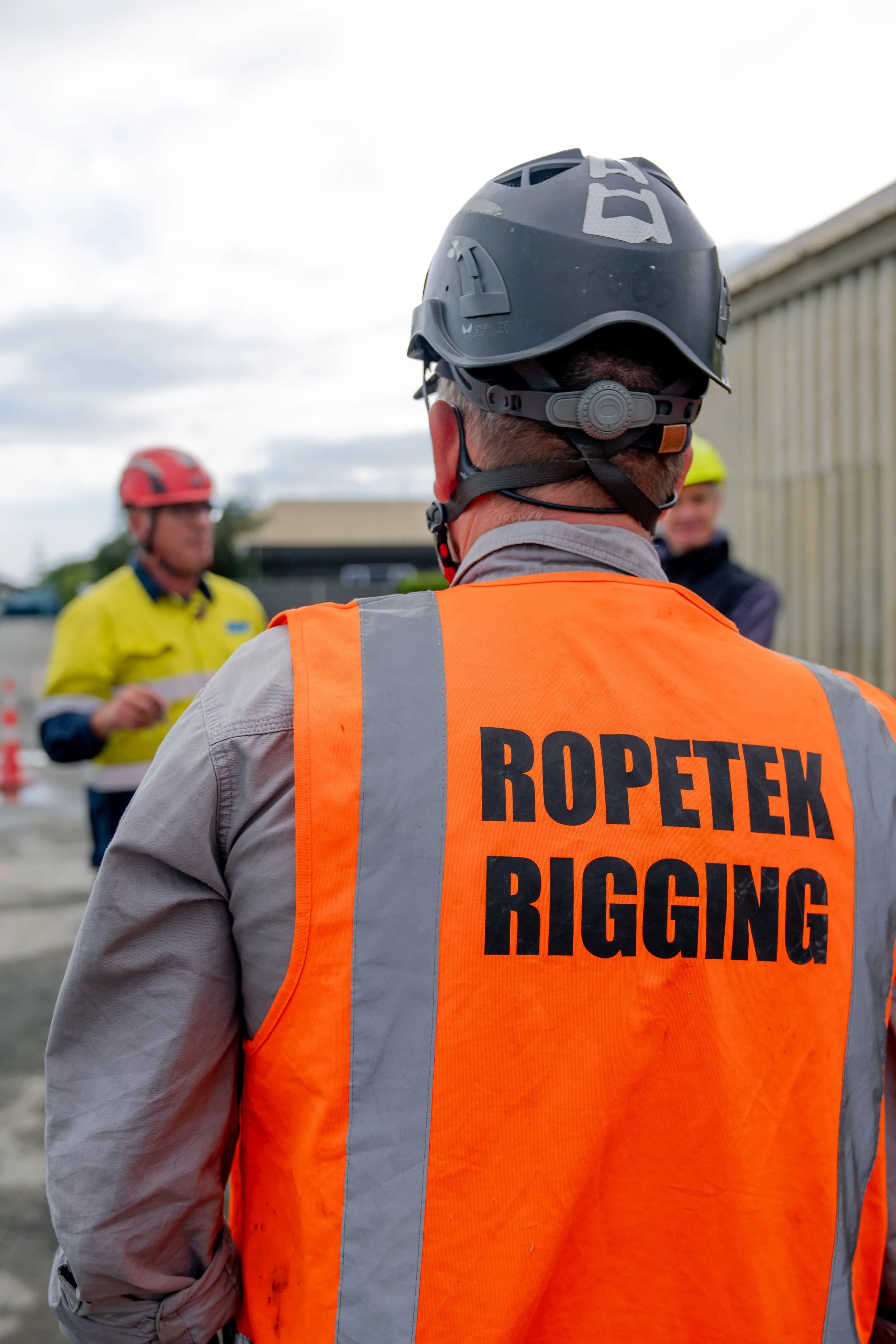 A construction worker wearing a helmet and an orange vest with 'ROPETEK RIGGING' printed on the back, standing outdoors and facing away from the camera, with two other workers in the background.