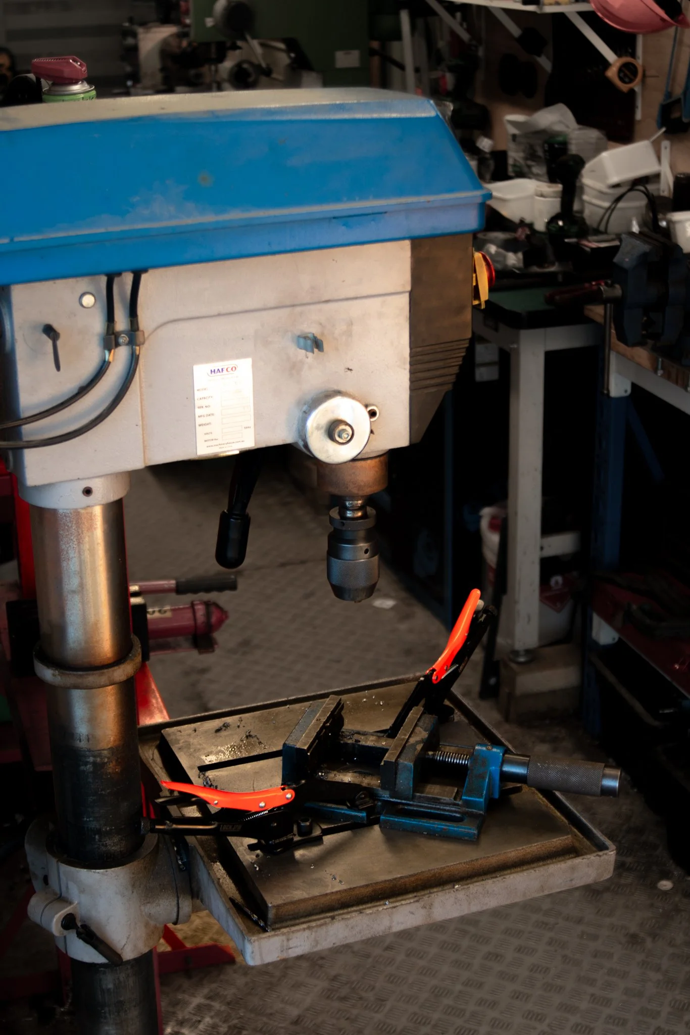 A drill press in a workshop with a clamp attached to the drill table.