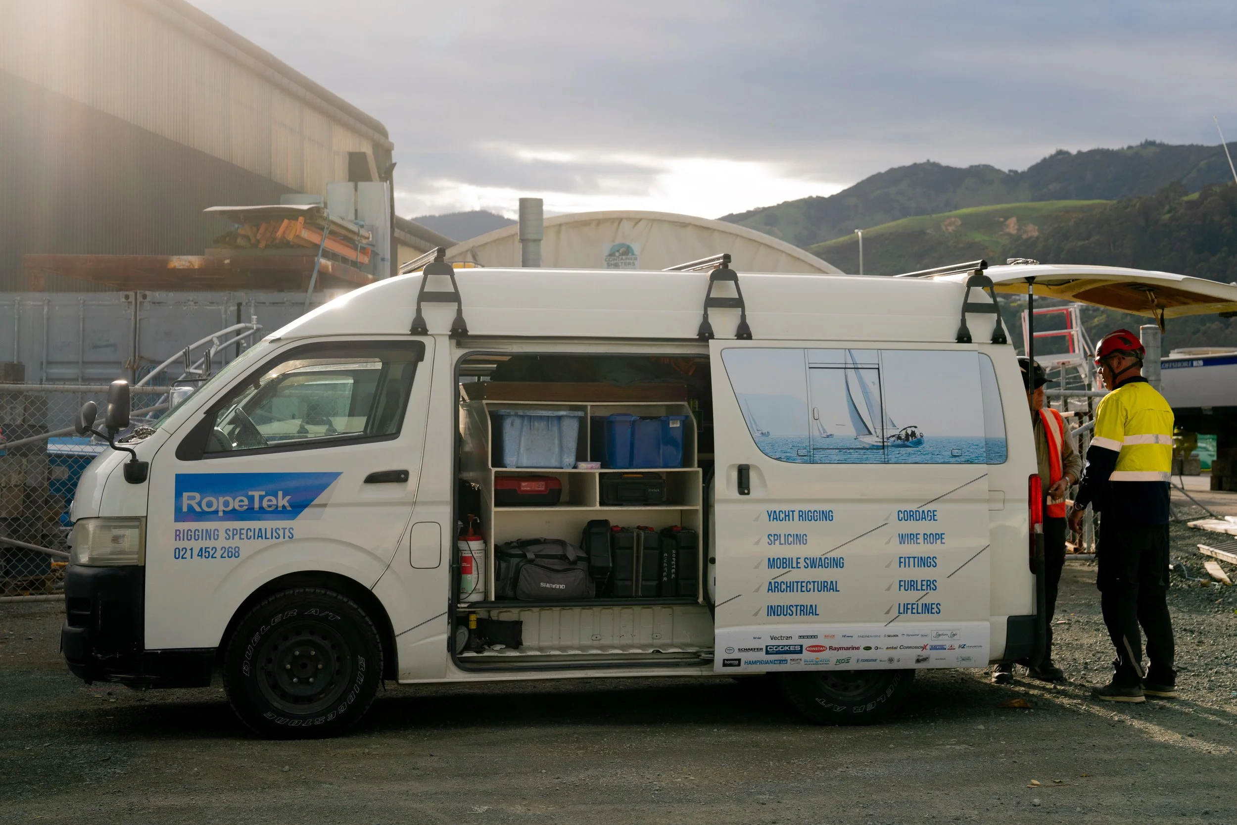 White van with open side door displaying equipment and side graphic, parked outdoors with two workers in safety gear talking beside it, boat and hills in background.