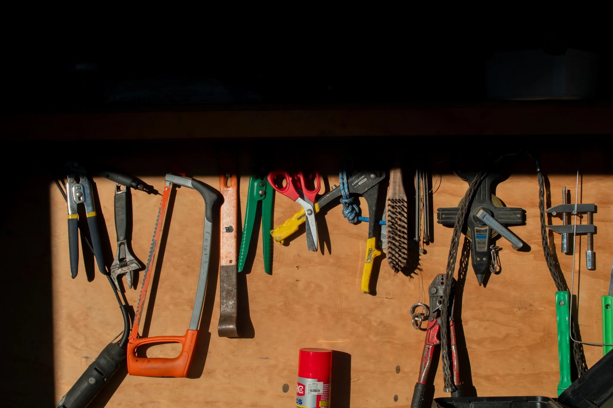 Tools hanging on a wooden wall, including a saw, pliers, scissors, paint scraper, clamp, and a spray can.