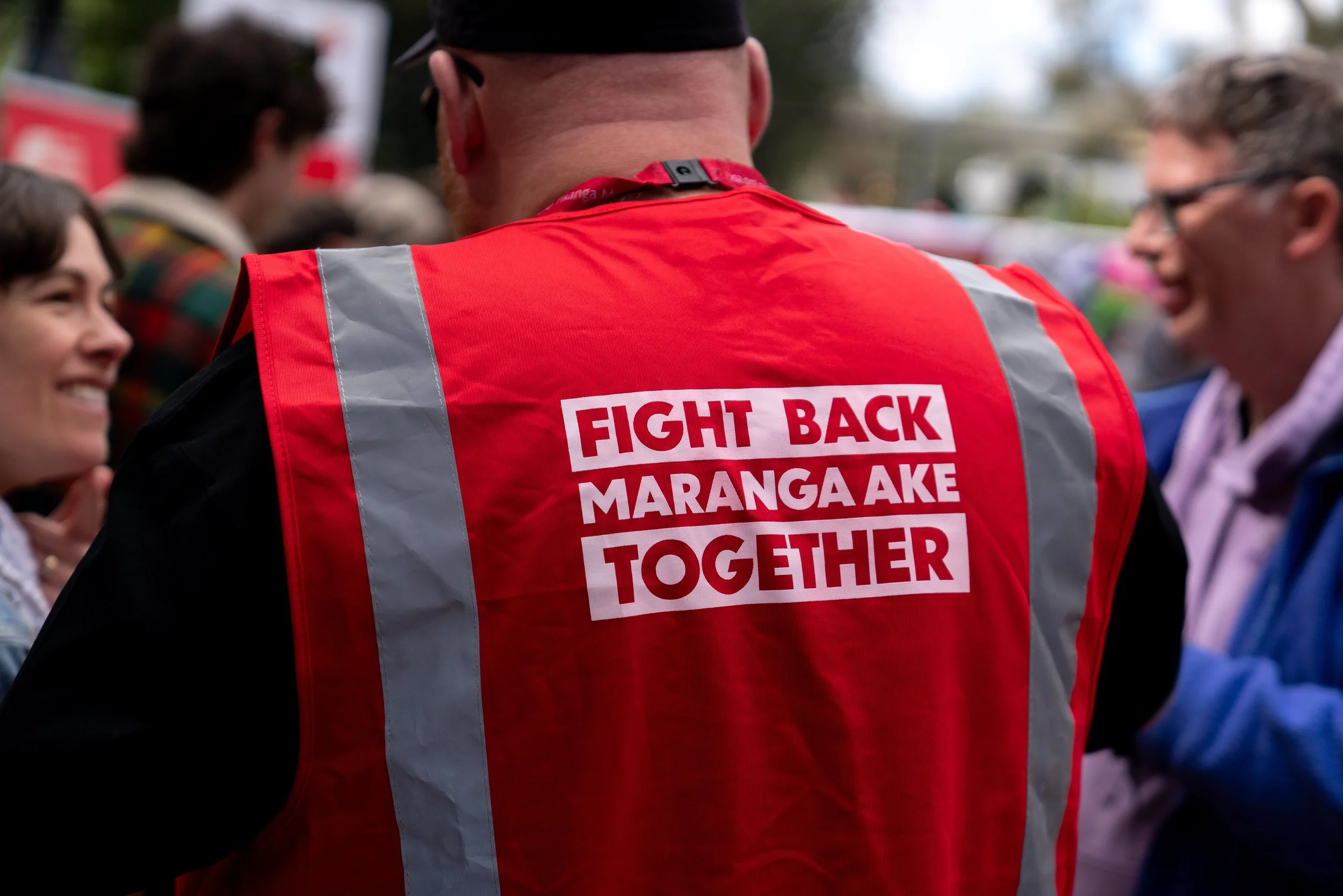 A person in a red safety vest with reflective stripes, with the message "FIGHT BACK MARANGA AKE TOGETHER" printed in white on the back, at a gathering with smiling people.