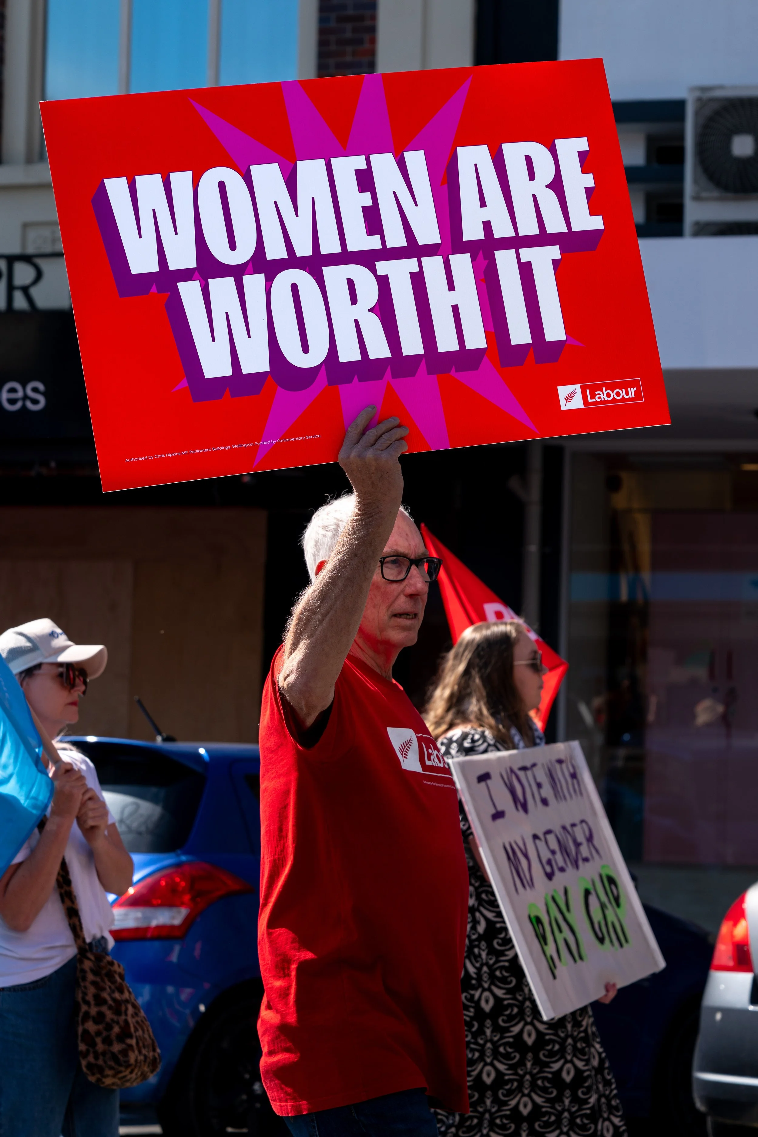 A person holding a sign that says 'Women are worth it' during a parade or protest, with other protesters holding signs in the background.