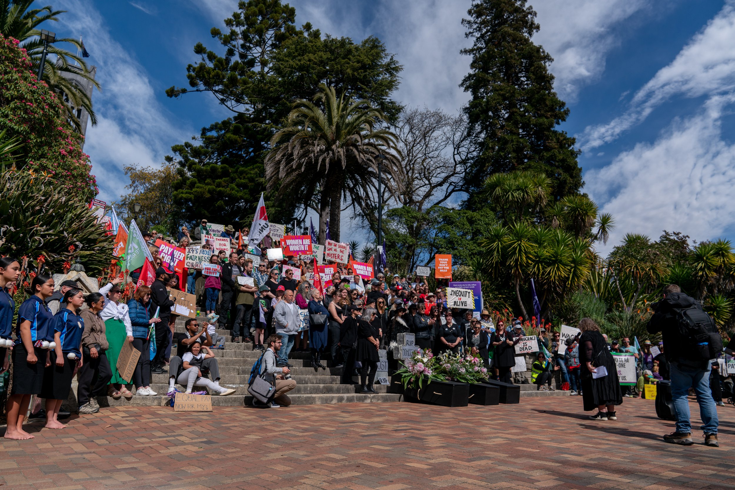 A large group of people gathered on steps outdoors during a protest, holding signs and flags advocating for women's rights and social justice, under a cloudy blue sky with lush green trees.