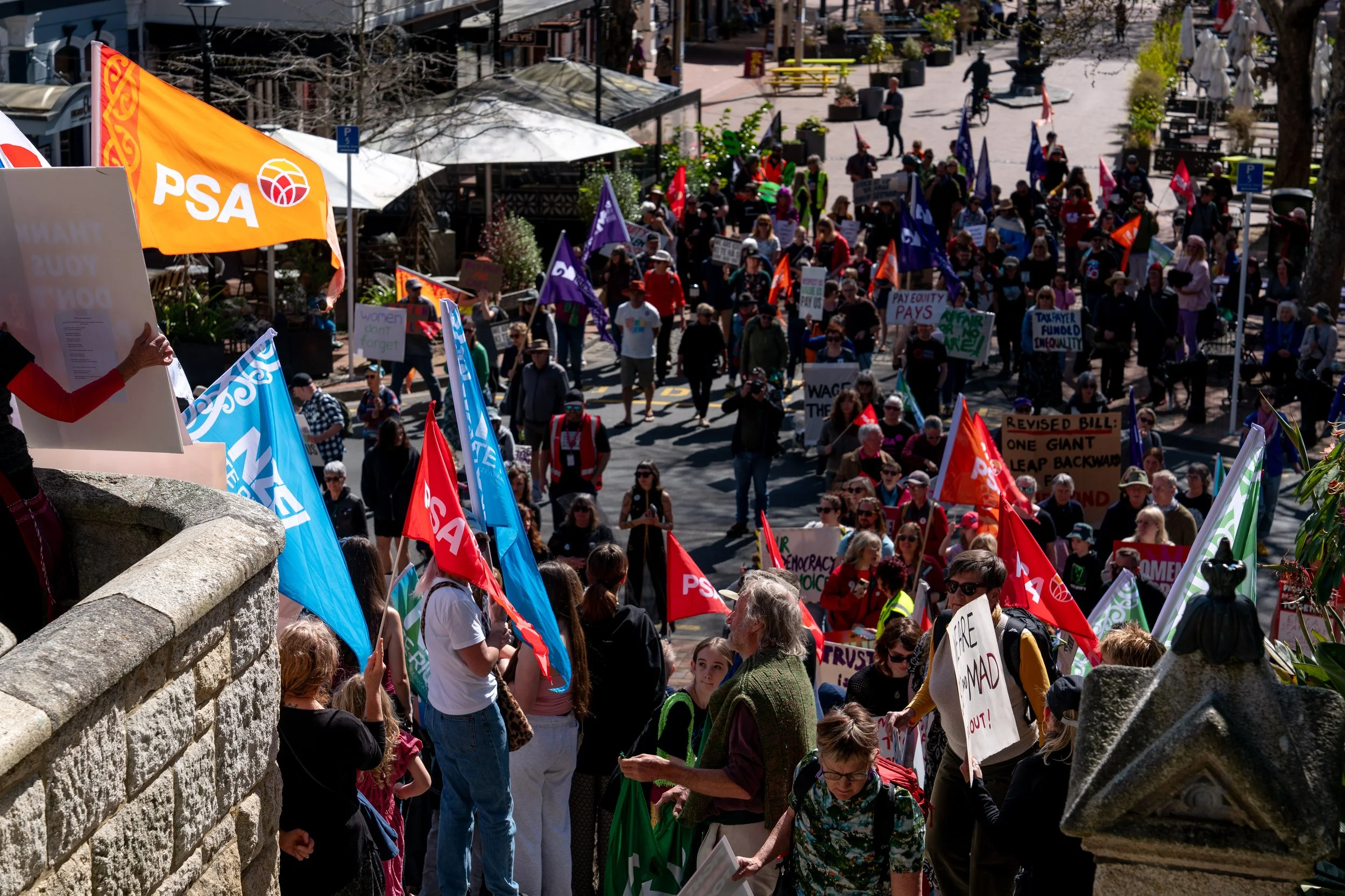 Crowd of people participating in a protest or march, holding various flags and signs, on a sunny day in an urban area with trees, outdoor seating, and buildings.