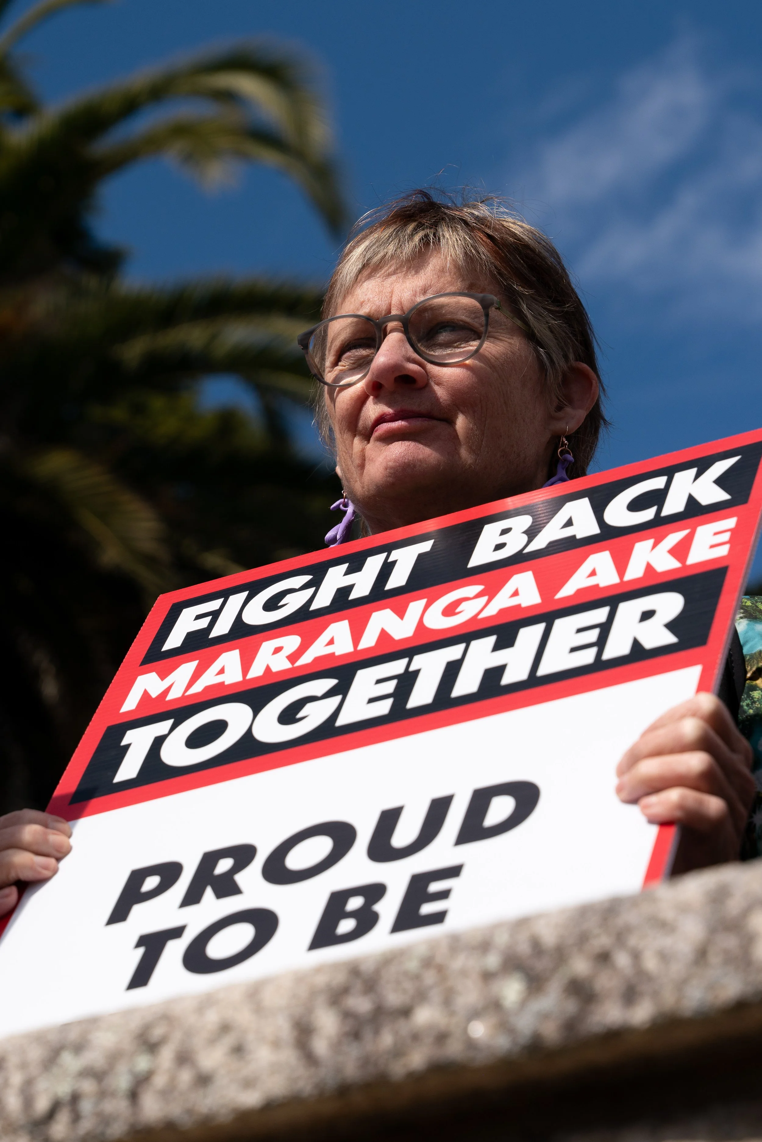 A woman with short gray hair and glasses holding a protest sign that reads 'Fight Back Maranga Ake Together' and 'Proud to Be' against a sunny sky with palm tree leaves in the background.