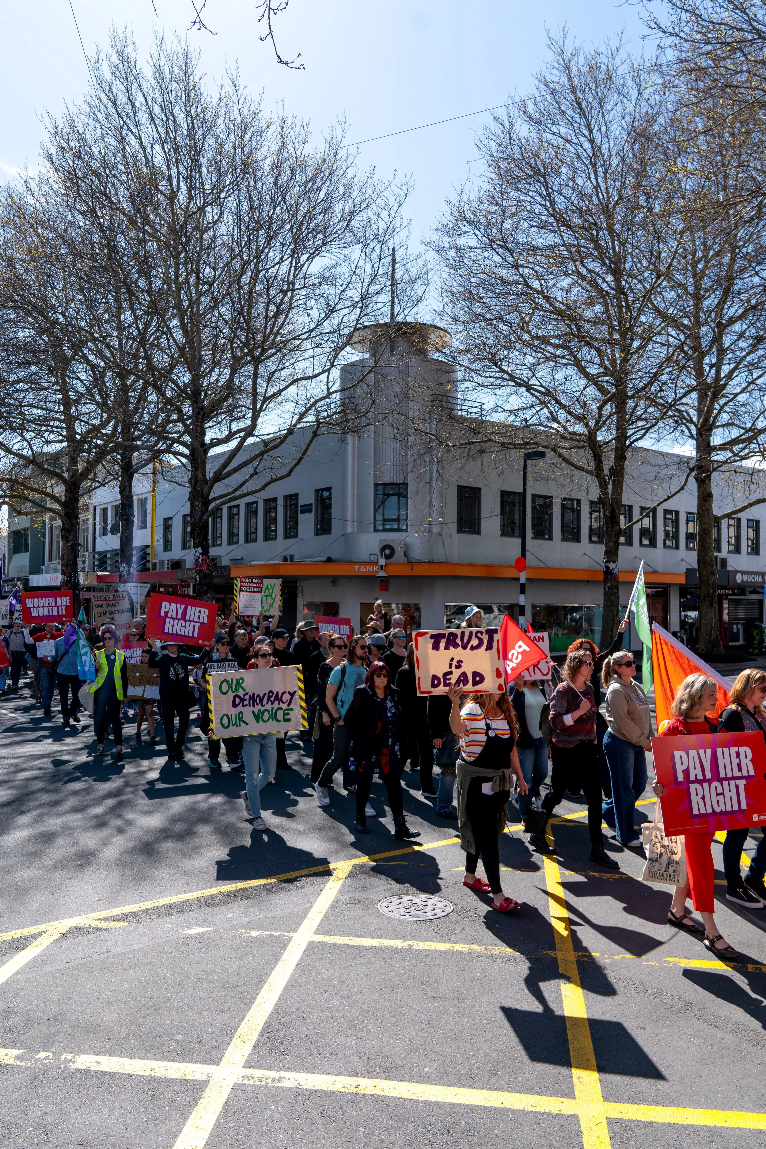 A group of protesters marching on a city street holding signs advocating for women's rights, democracy, and justice, with some signs reading "PAY HER RIGHT," "WOMEN ARE WORTH IT," "OUR DEMOCRACY OUR VOICE," and "TRUST IS DEAD." The protesters are wal