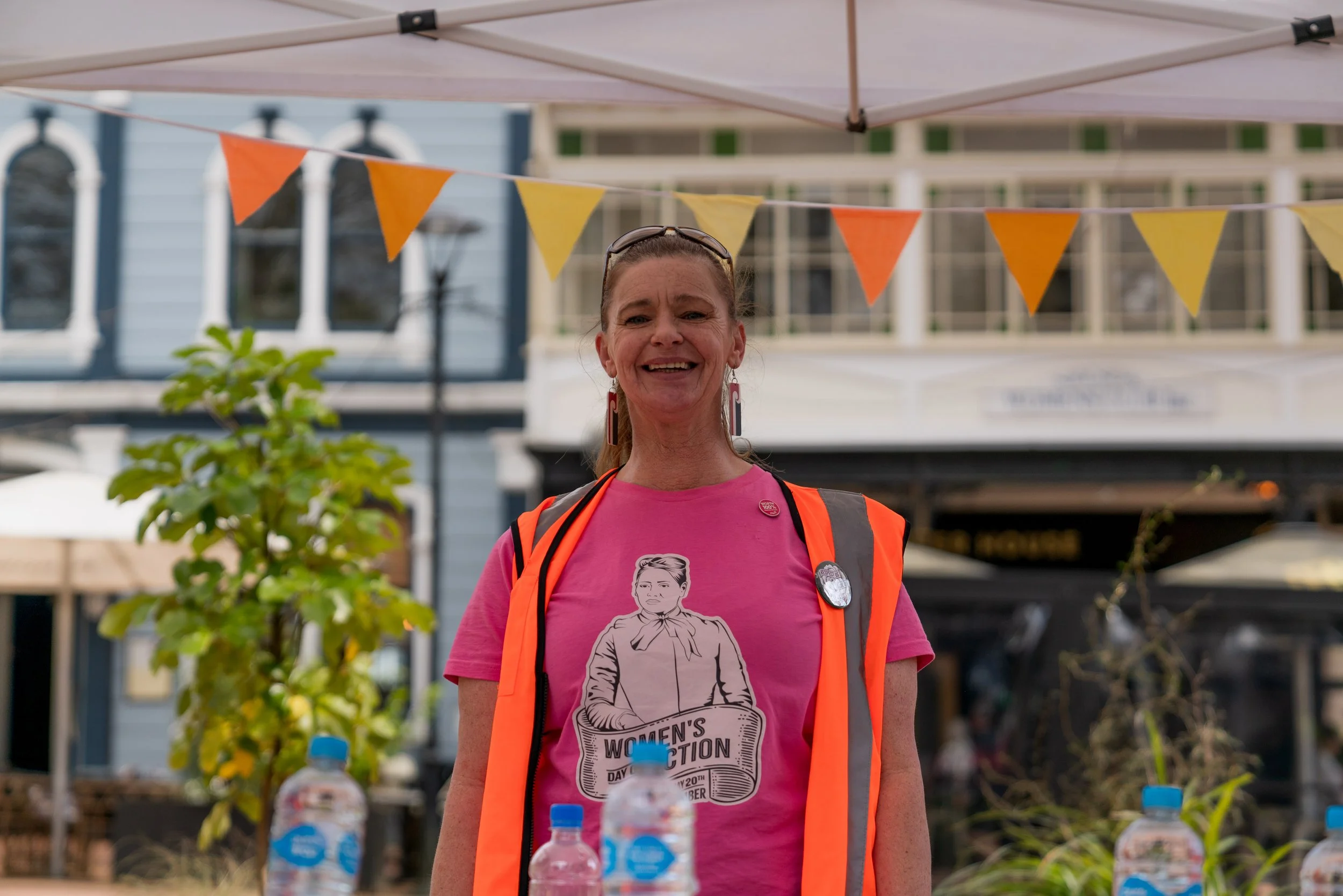 A woman smiling, wearing a pink t-shirt with a woman illustration and the words 'Women's Action Day October', and an orange safety vest. Behind her are a table with water bottles, outdoor seating, and a building with banners and bunting flags.