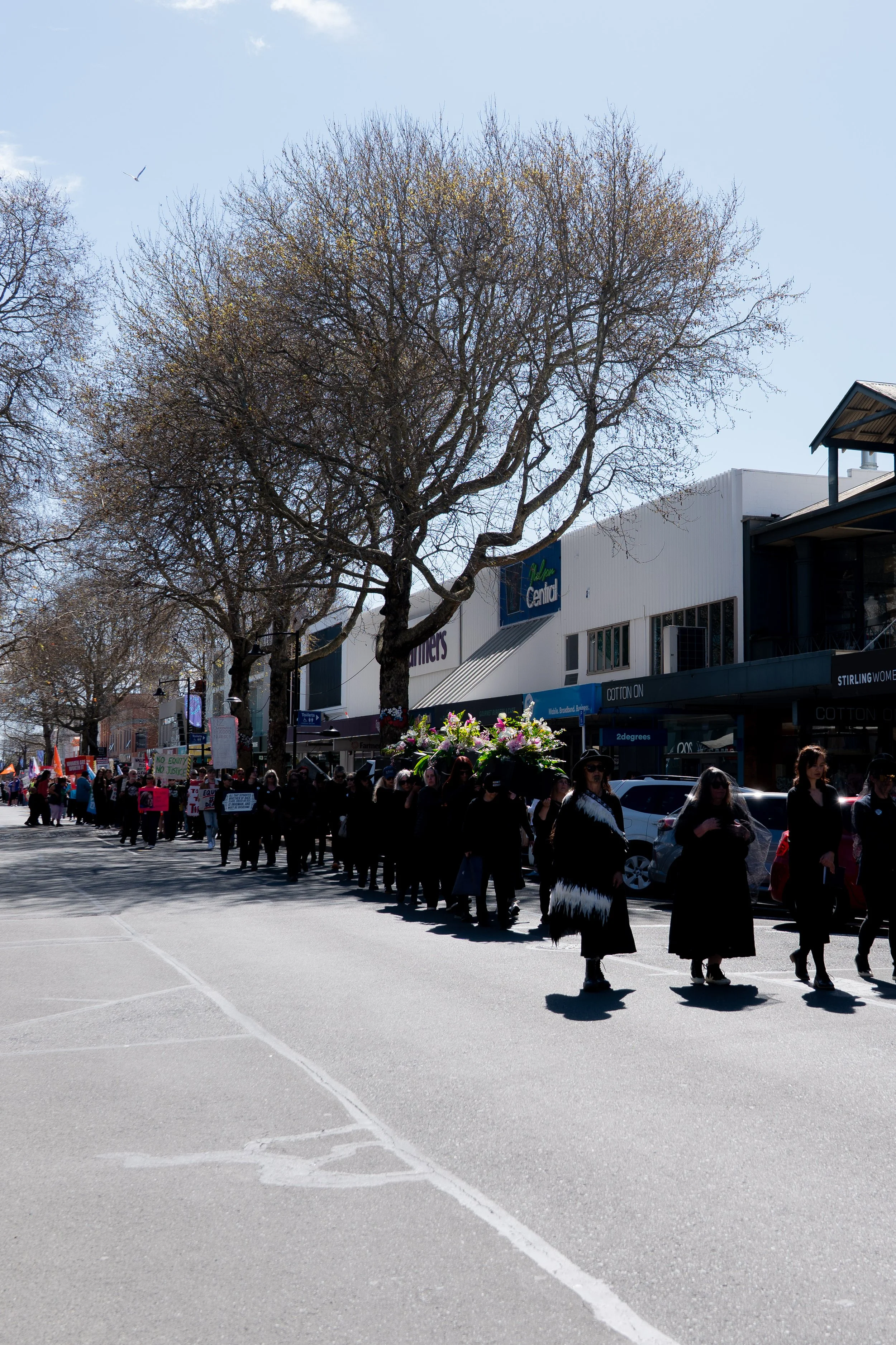 A funeral procession with people walking in mourning down a city street, carrying a casket adorned with flowers.