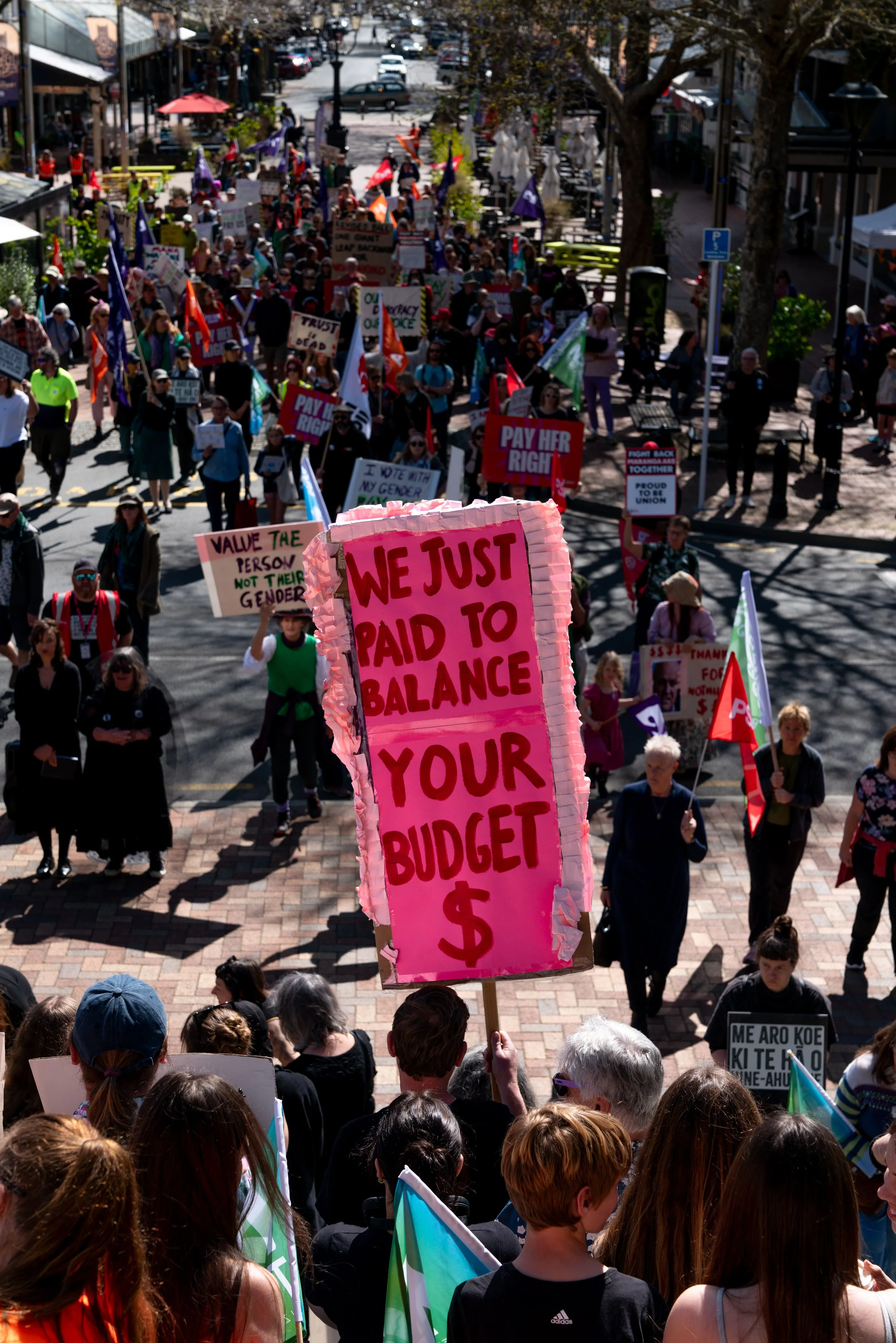 A large crowd of protesters marching down a city street, holding various signs and flags. One prominent pink sign reads, 'WE JUST PAID TO BALANCE YOUR BUDGET $.'