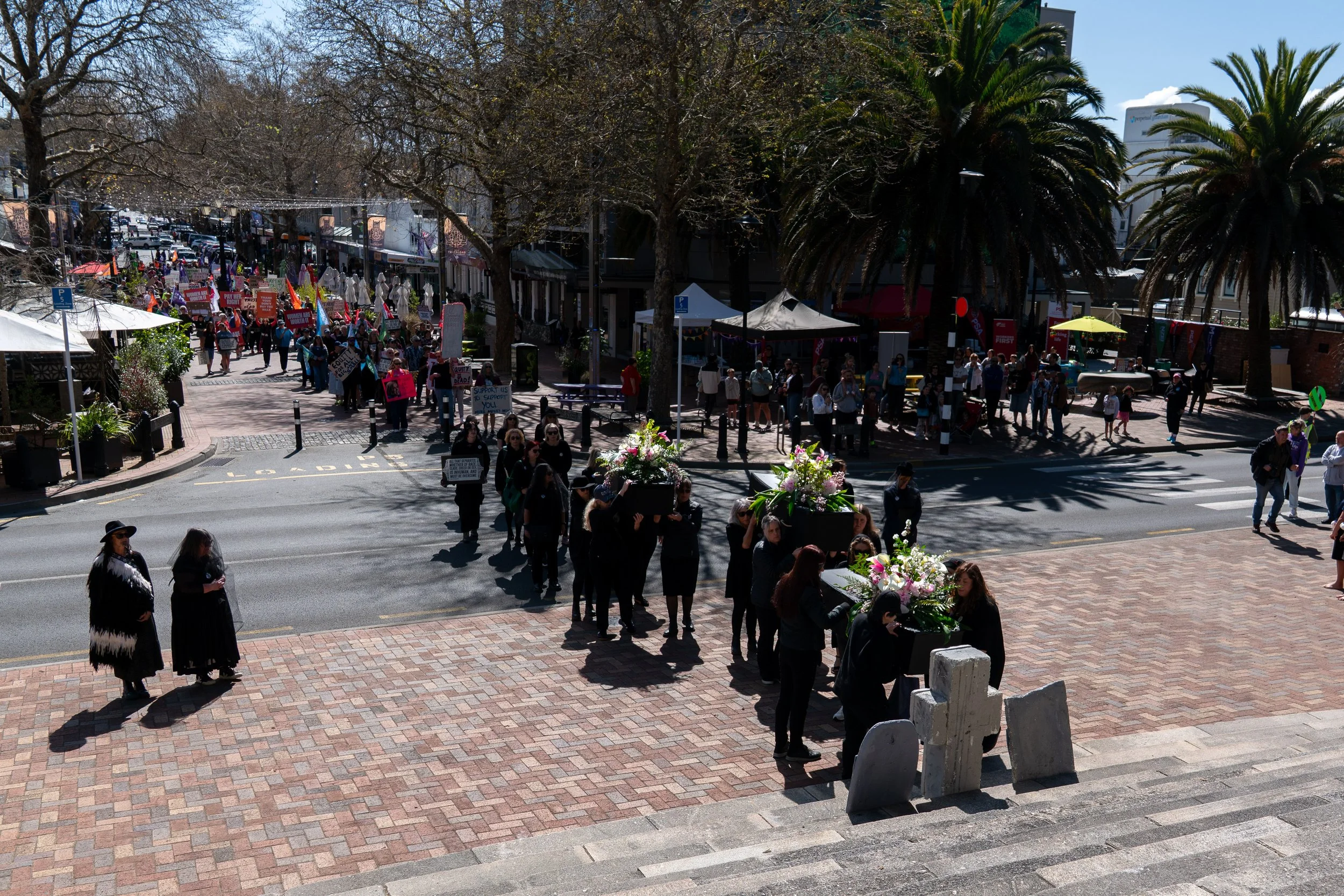 A street protest or march with many participants holding signs and banners, some wearing black. The group is walking along the road and over a crosswalk, with some carrying large flower arrangements. There are trees, tents, and shops in the backgroun