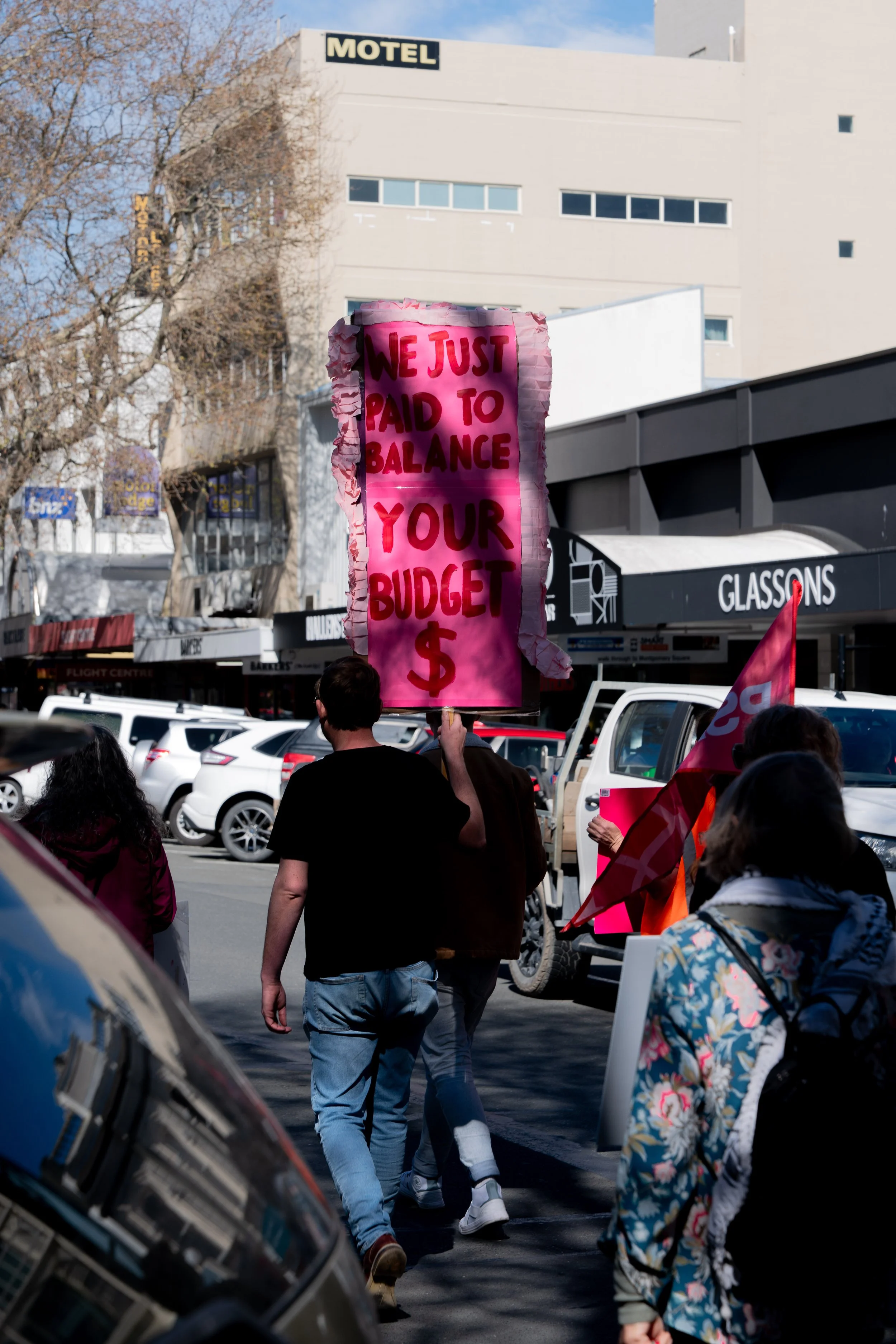 Protesters marching on the street with signs, including a large pink sign with red letters that says, 'We just paid to balance your budget $', during a demonstration.
