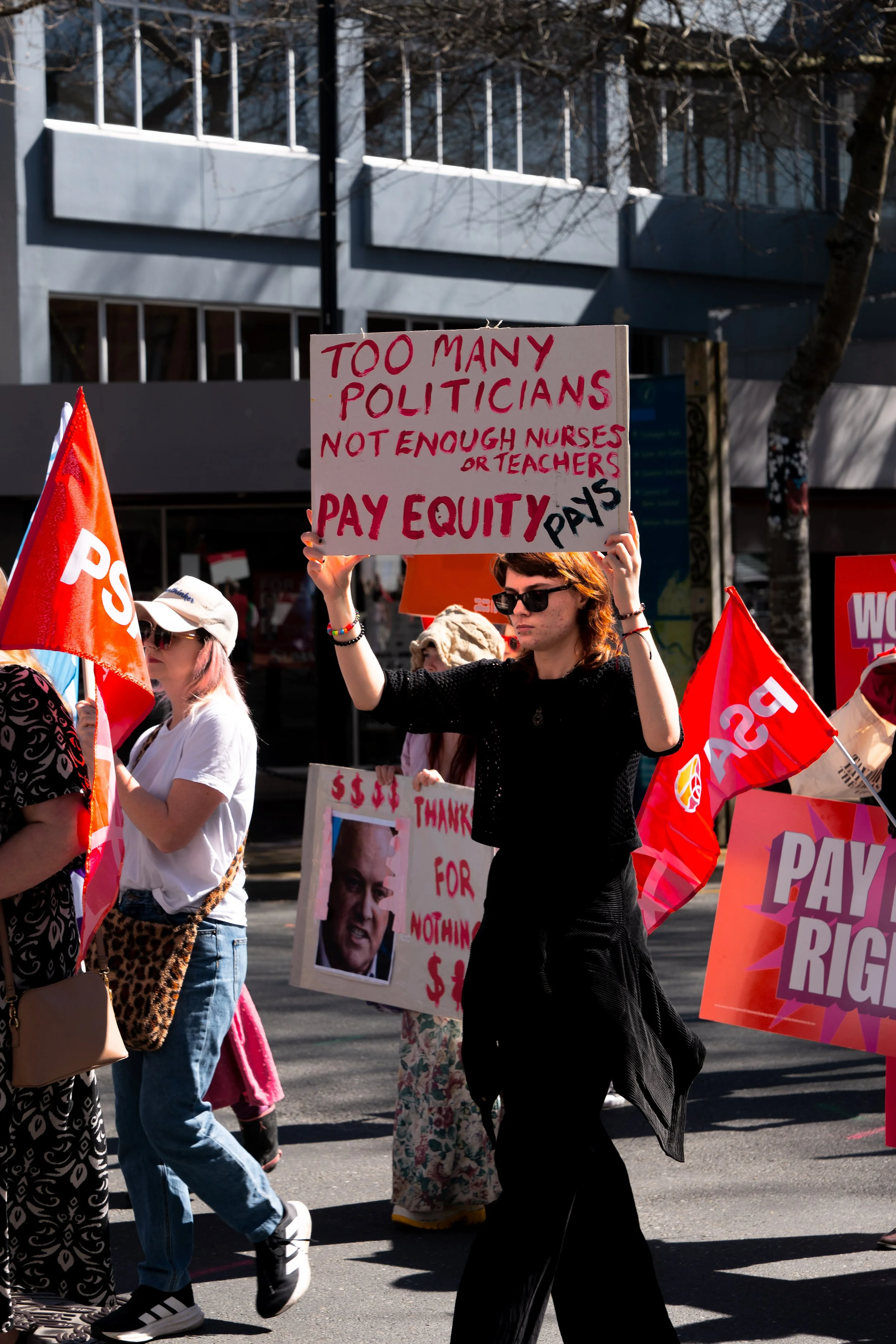 A woman holding a protest sign that reads 'Too many politicians, not enough nurses or teachers. Pay equity pays.' at a rally, surrounded by other protesters, some holding flags and signs.