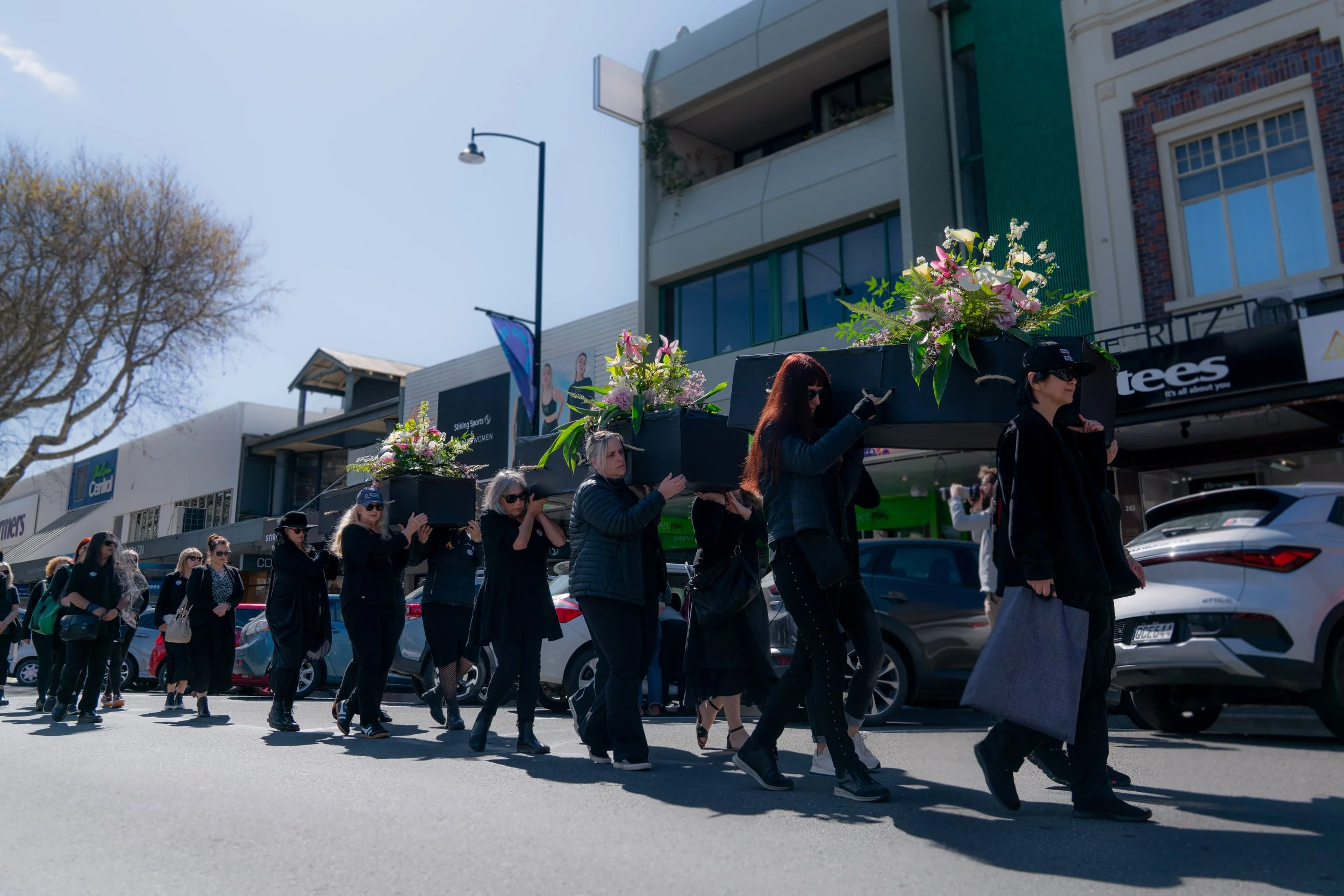 A procession of people walking on a city street, carrying a large black casket decorated with pink and white flowers.