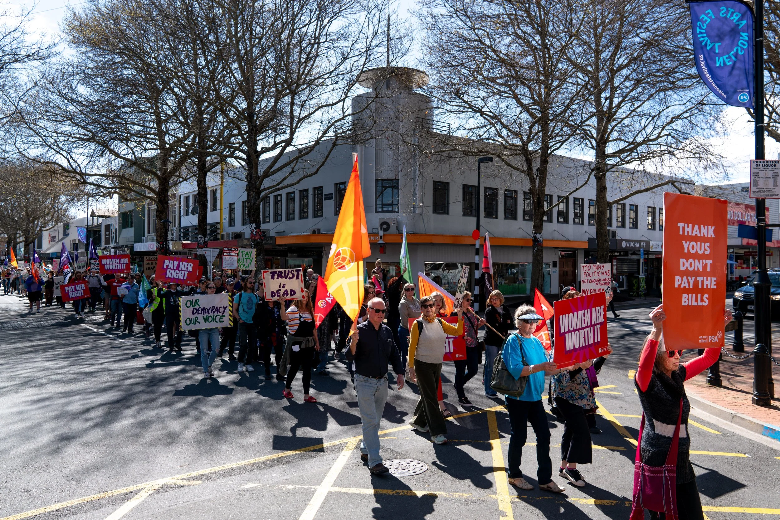 A large group of people participating in a protest march on a city street, holding signs advocating for women's rights, democracy, and against political bills. Some signs say 'Women Are Worth It,' 'Pay Her Right,' and 'Thank You Don’t Pay The Bills.'