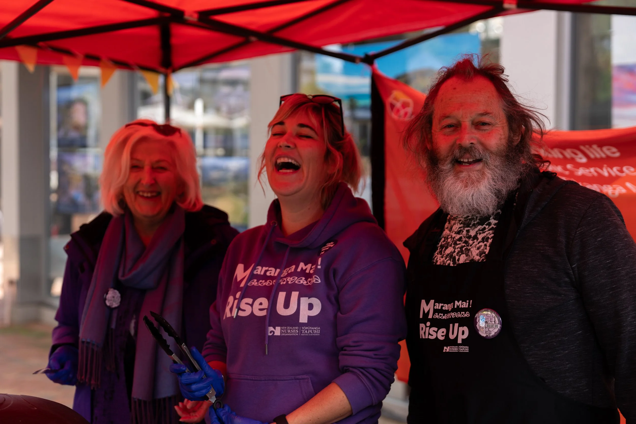 Three people smiling and laughing at an outdoor event under a red canopy, wearing purple and black clothing with 'Māori Mai! Rise Up' slogans, one person holding tongs, with banners and buildings in the background.