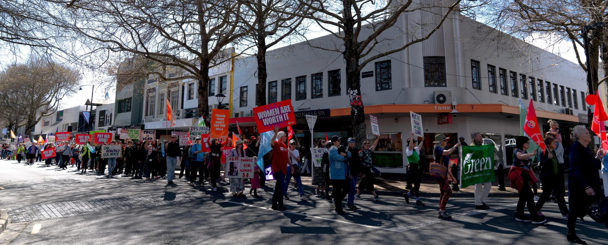 A large group of people marching in a protest or rally on a city street, holding signs with messages about women's rights, equality, and political issues. The march is taking place during daytime with leafless trees and buildings in the background.