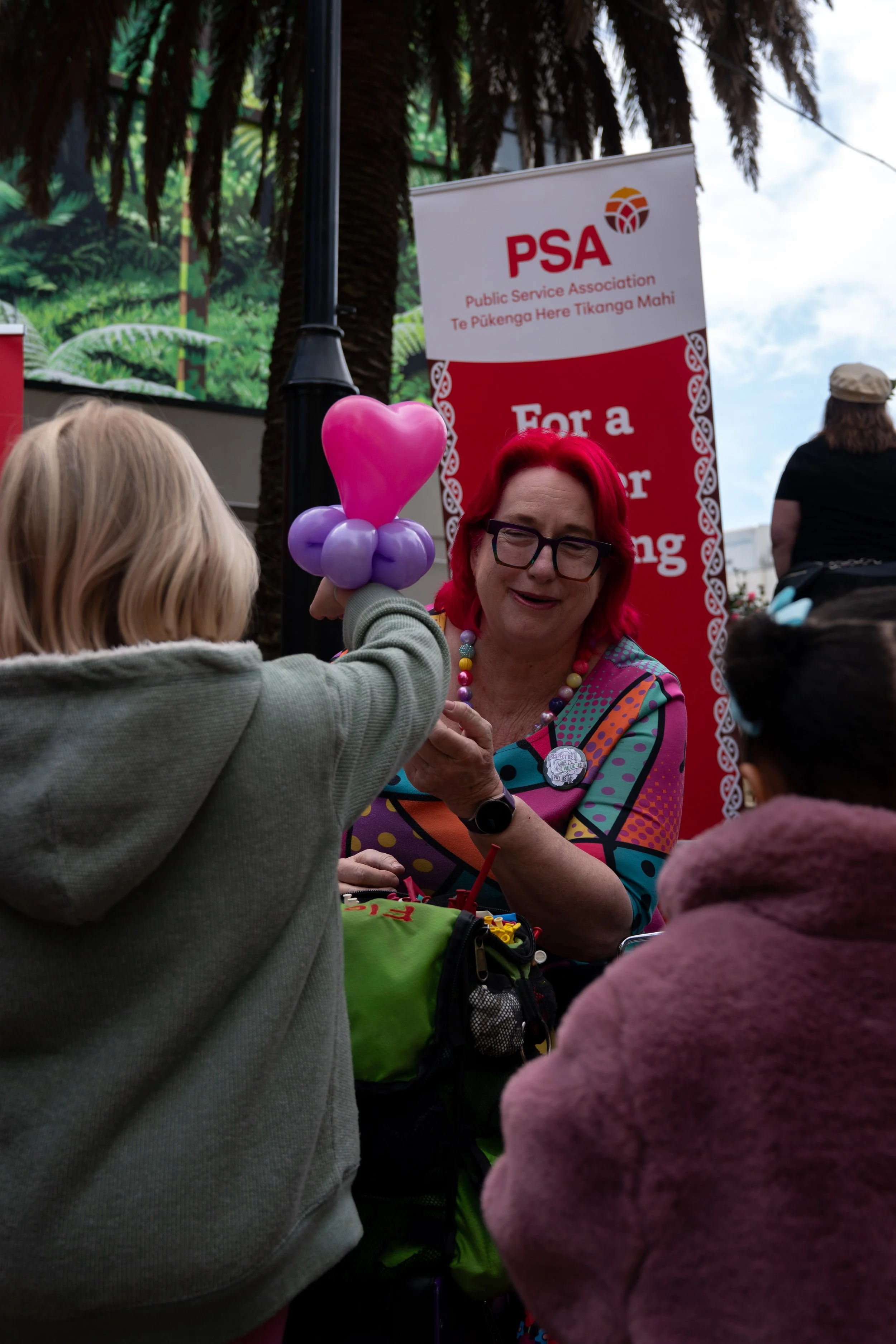 Woman with bright red hair and glasses smiling while talking to children, one of whom is holding a pink balloon shaped like a heart, at a public event with a red PSA banner in the background.