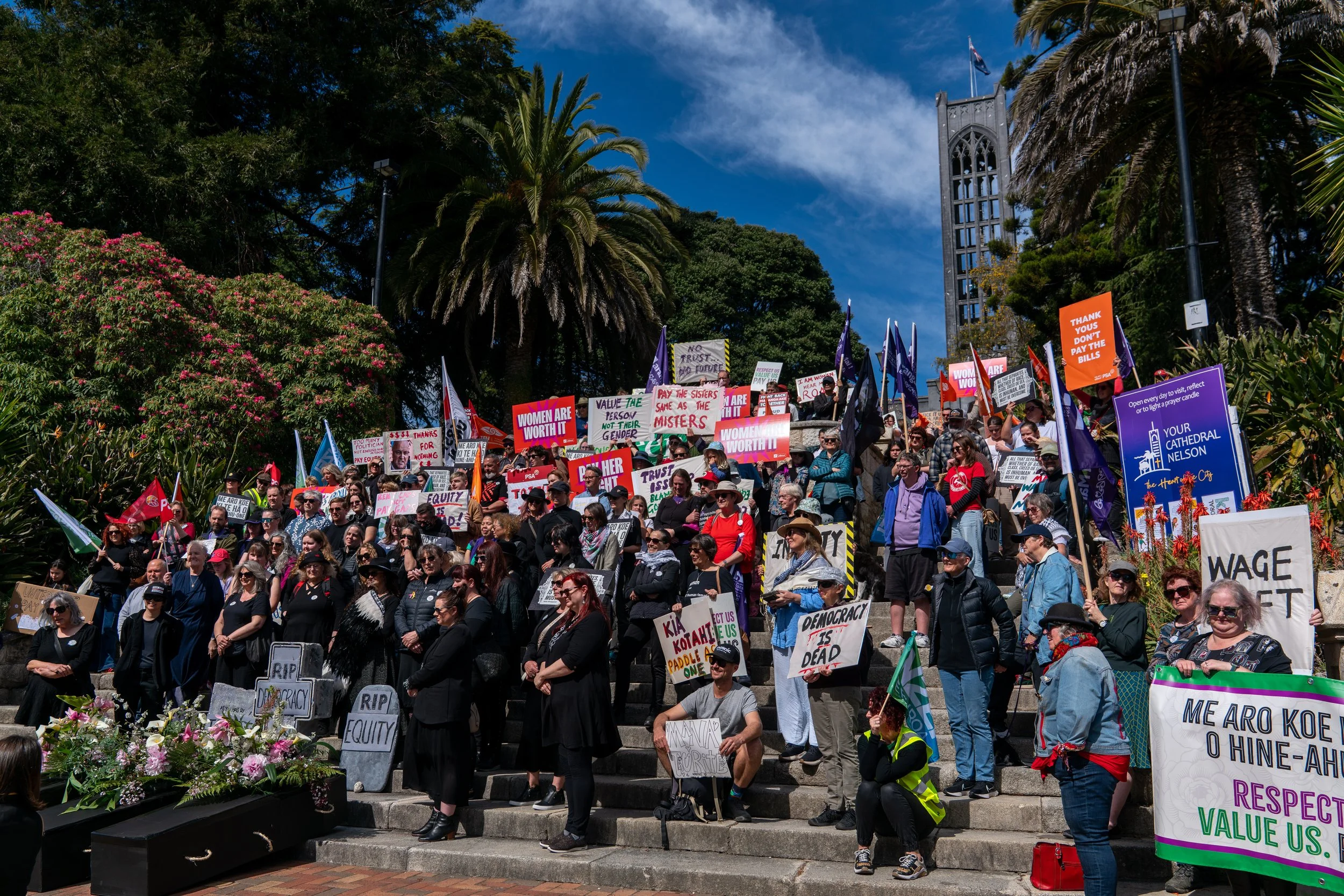 A large crowd of protesters gathered on outdoor steps, holding signs and banners advocating for women's rights and justice, with trees and a tall building in the background under a blue sky.