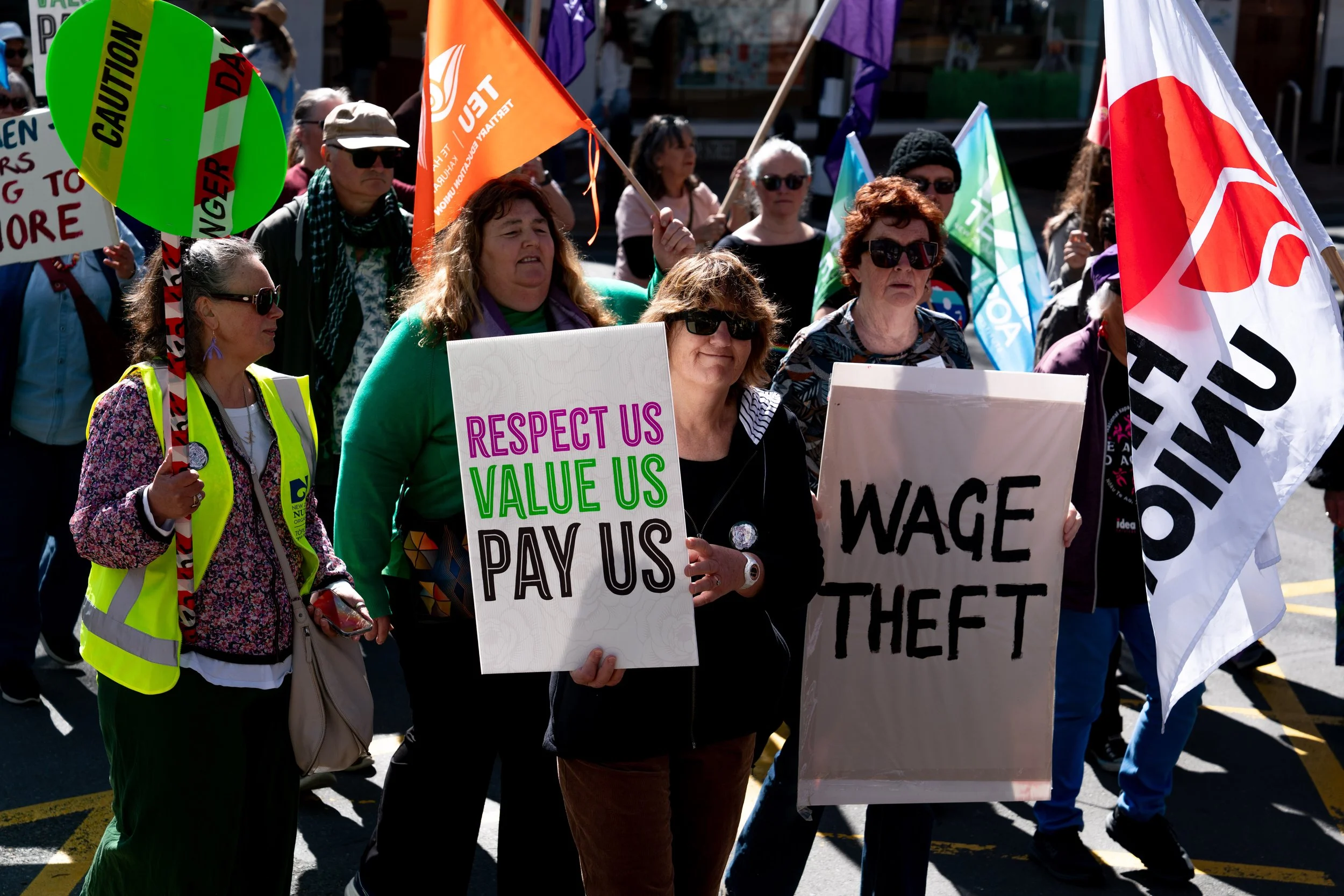 Group of people participating in a protest march, holding signs and flags advocating for workers' rights and fair wages, including messages like 'Respect Us Value Us Pay Us' and 'Wage Theft'.