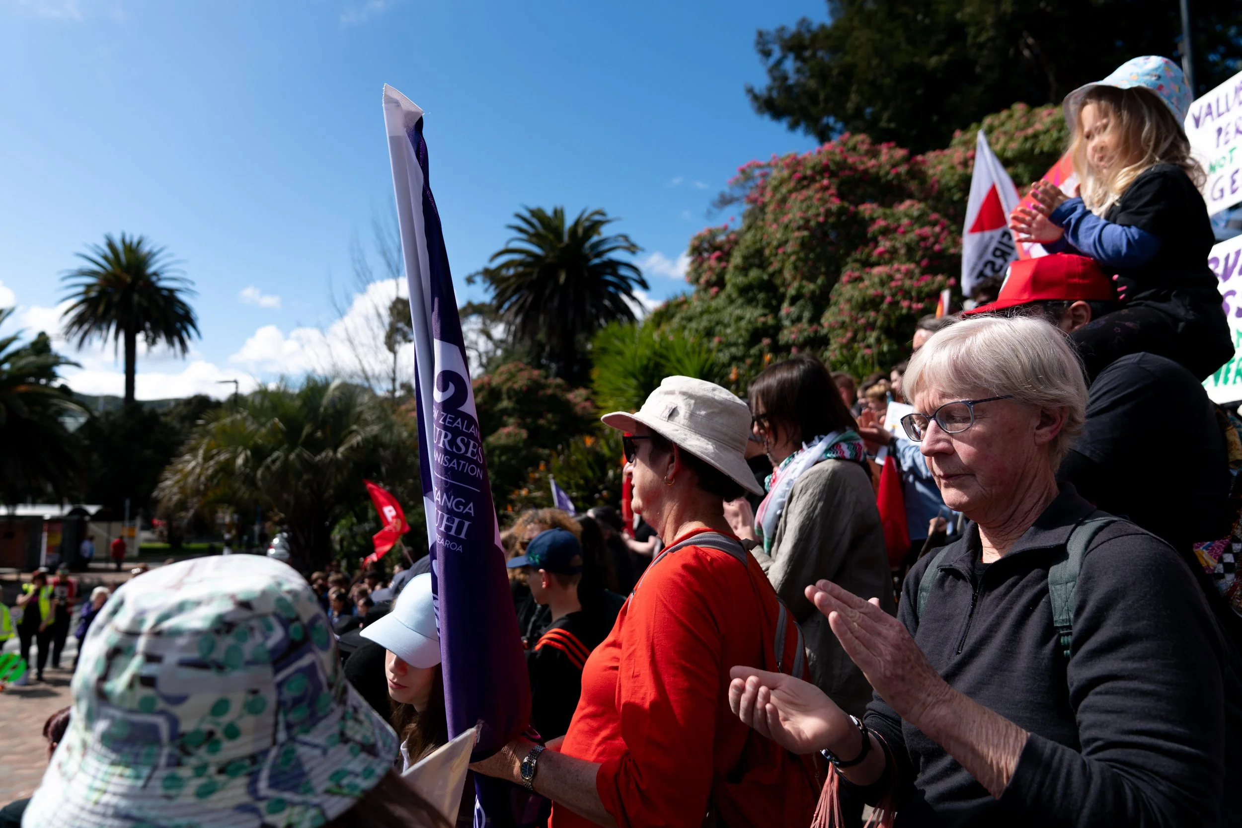 Group of people participating in a protest or rally outdoors. Some are praying or with hands clasped, holding flags and signs. The setting includes palm trees, flowering bushes, and a clear blue sky.