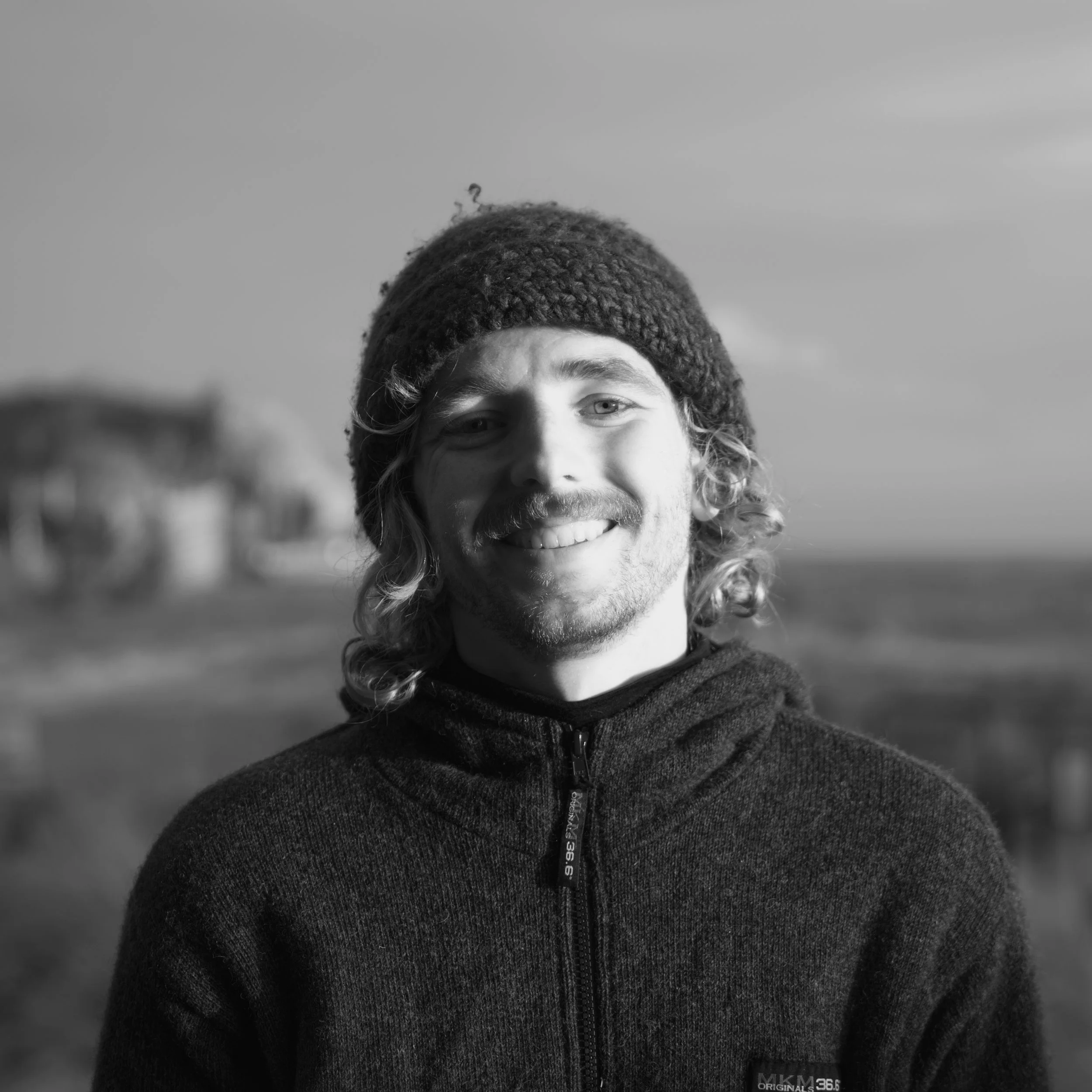Black and white photo of a smiling man with long, wavy hair, wearing a wool hat and a zip-up sweater, outdoors with a building and sky in the background.