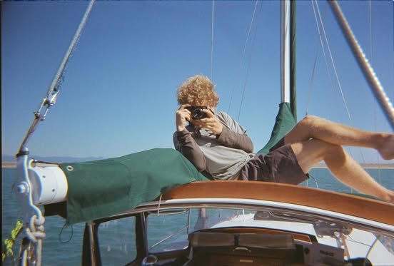 Person with curly hair taking a photo while lying on the sun deck of a sailboat, with water and sky in the background.