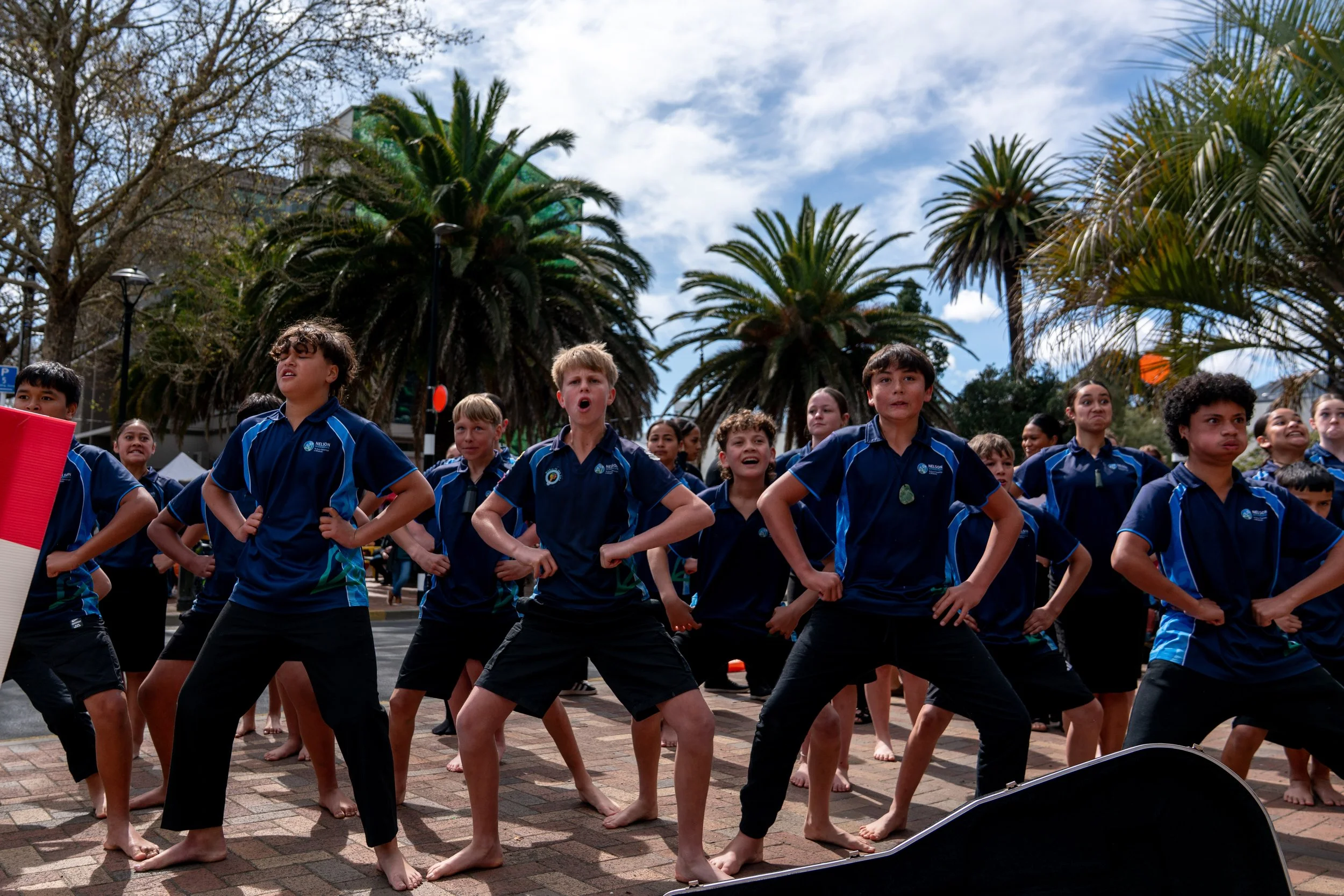 Group of young students in matching blue shirts and black pants performing a choreographed dance or exercise outdoors on pavement, with palm trees and a partly cloudy sky in the background.