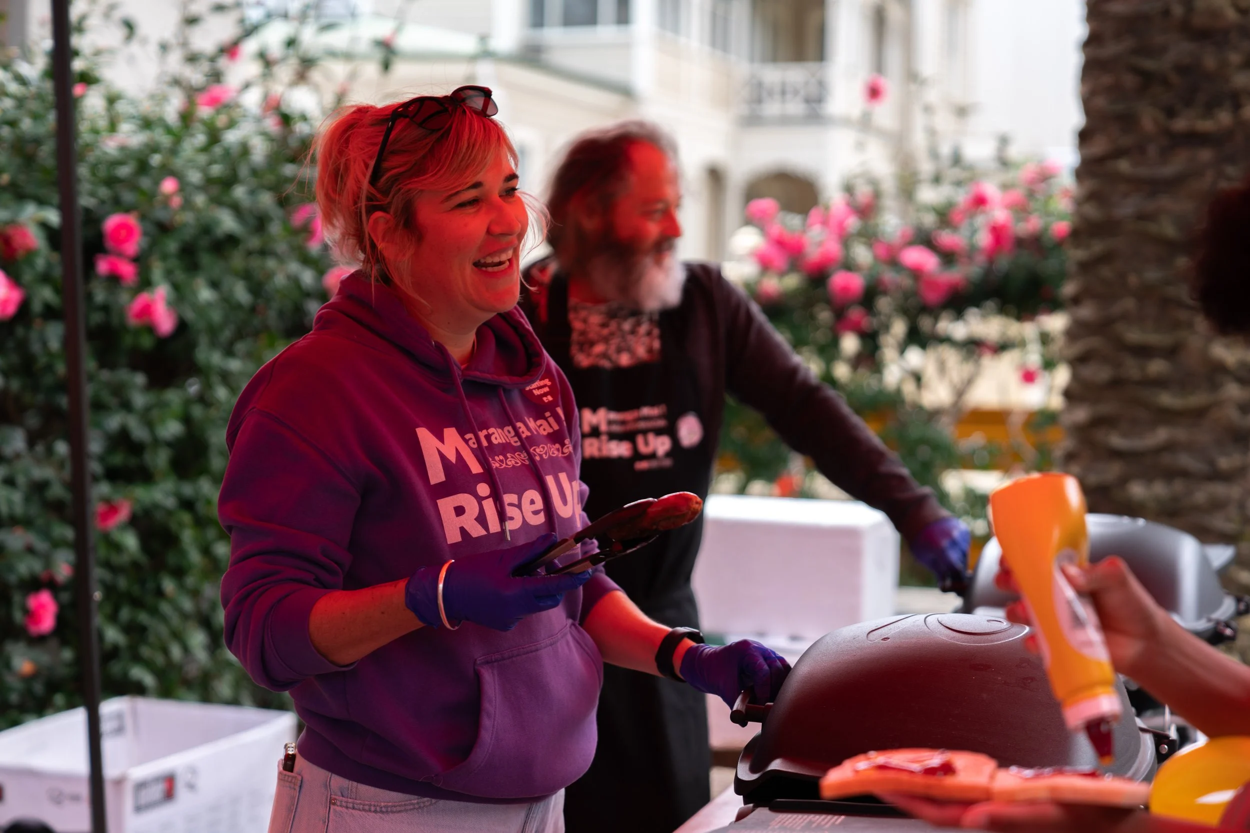 People serving food at an outdoor event with pink flowers in the background.
