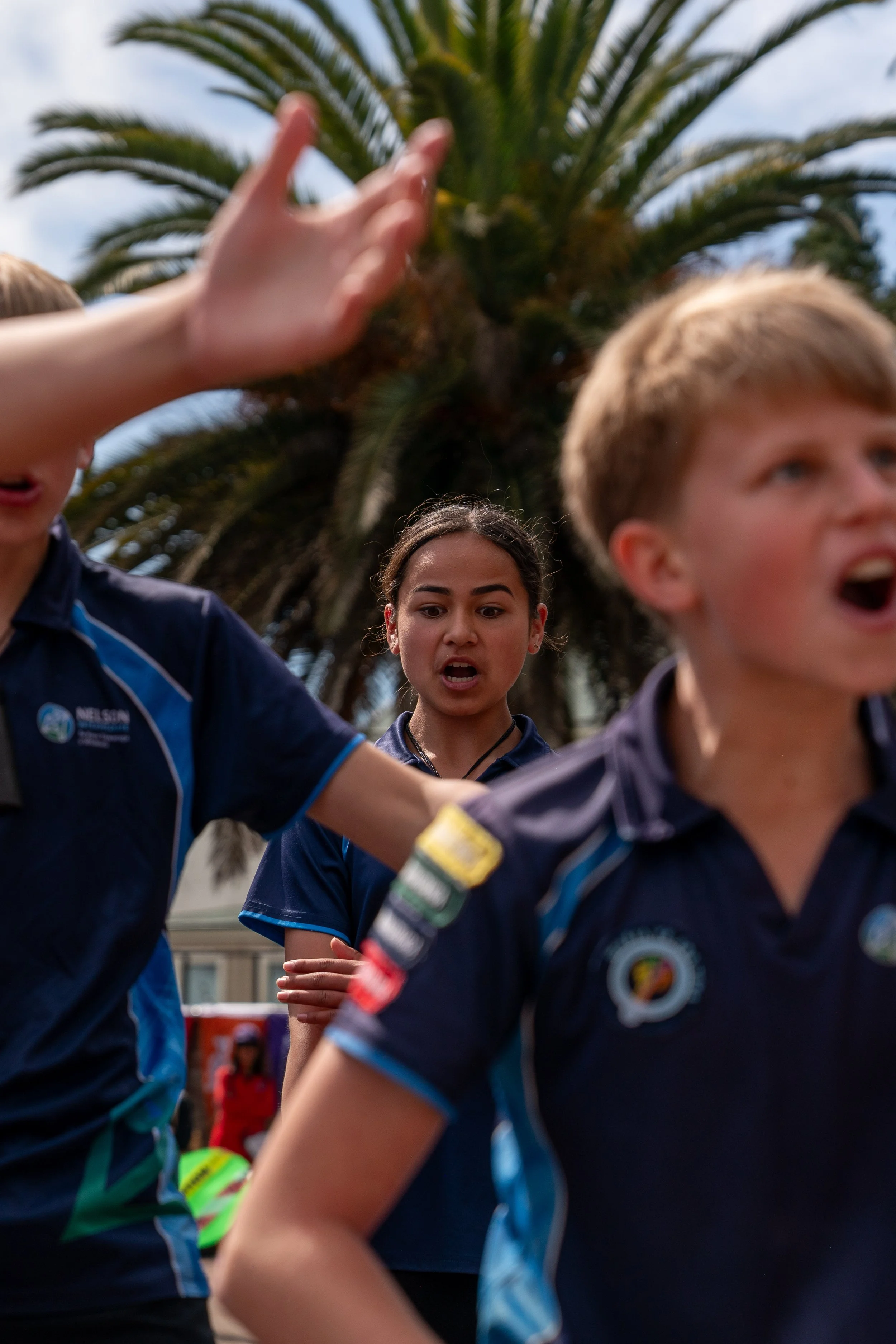 Children in sports uniforms outdoors, with palm trees and a building in the background, appearing to be in a discussion or argument.