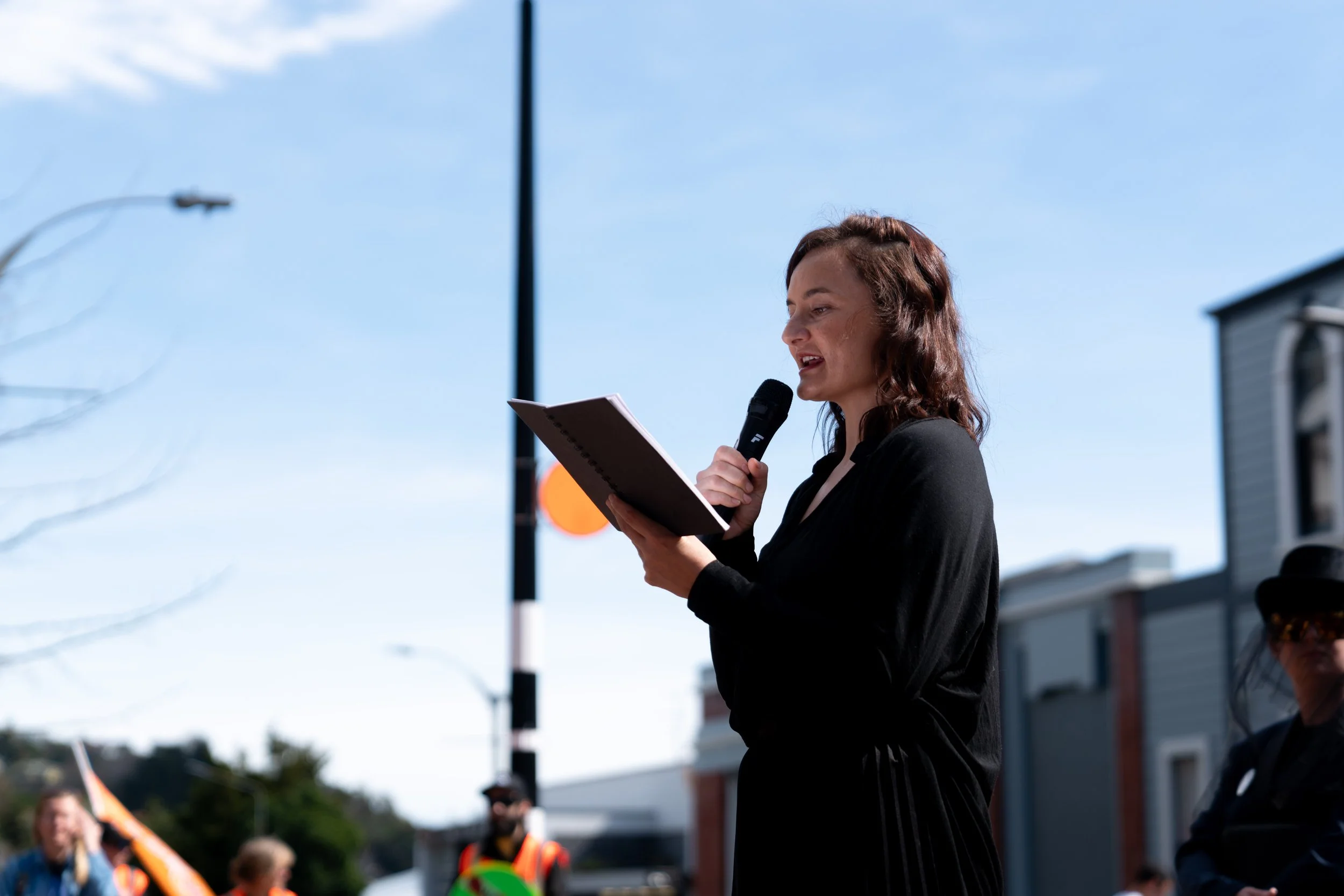 Woman speaking into a microphone while reading from a notebook at an outdoor event on a sunny day.