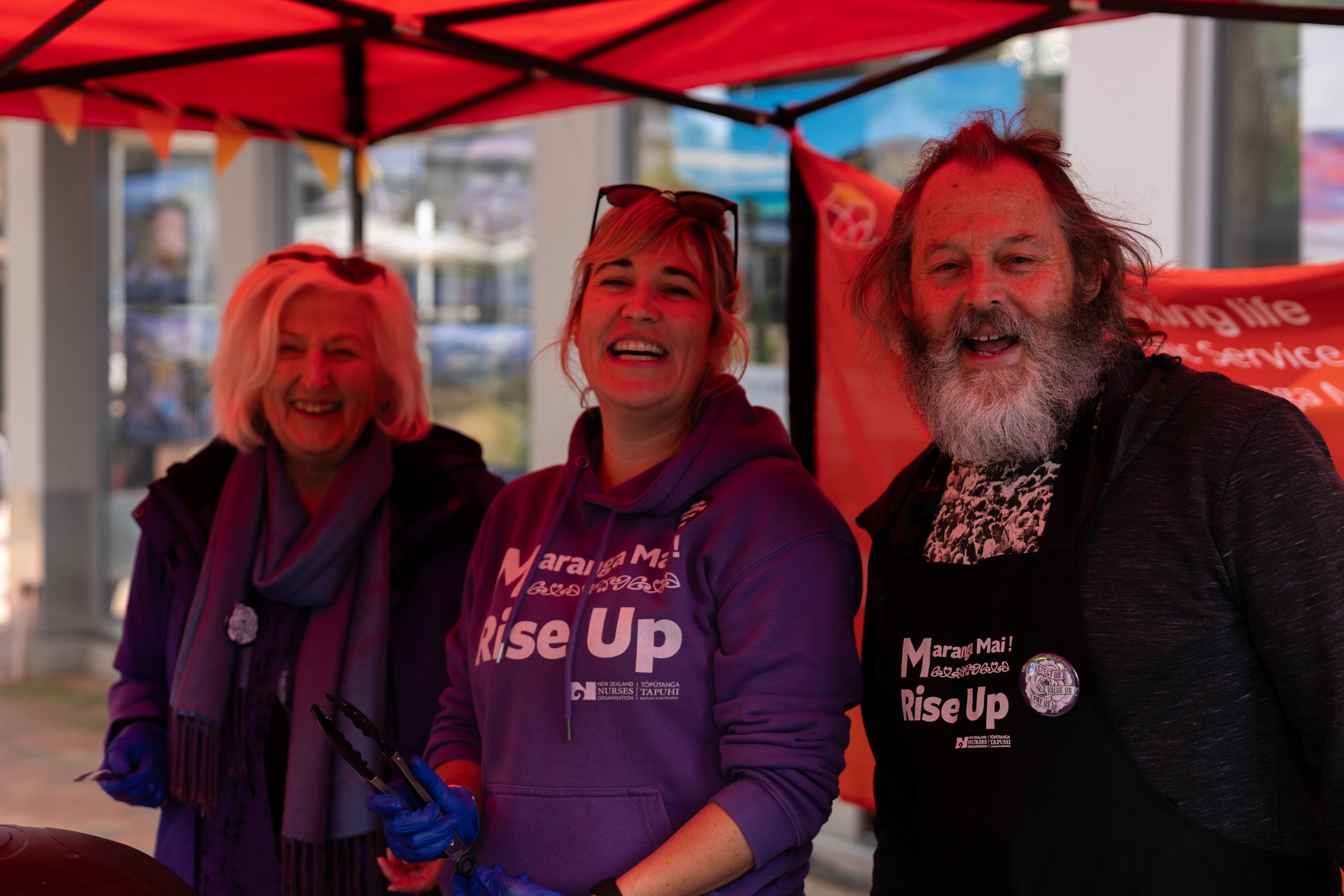 Three smiling people at a community event, standing under a red canopy, wearing Marama Mai! Rise Up shirts, with one woman holding tongs and wearing gloves.