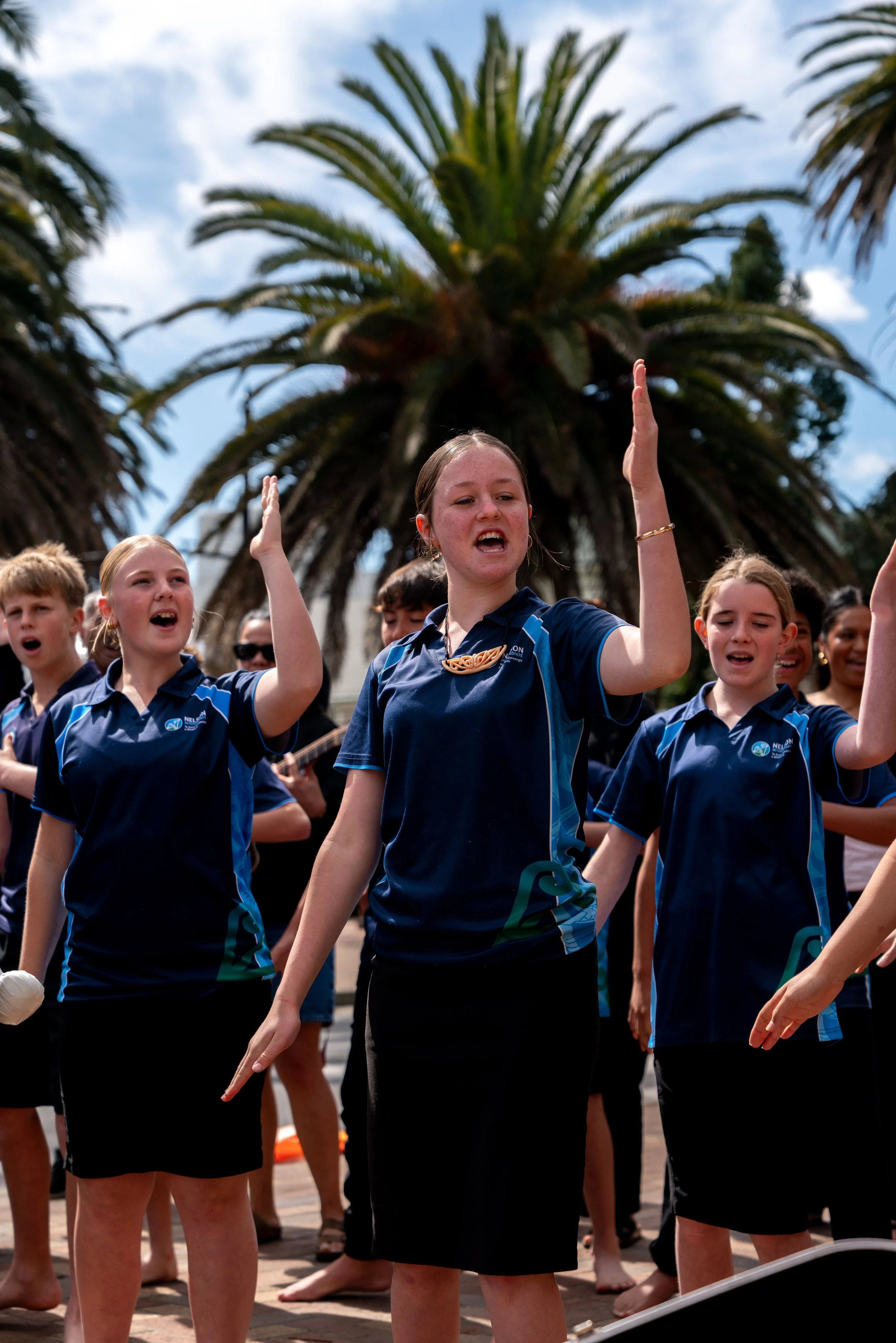 Group of young girls in blue shirts performing outdoors with palm trees in the background.