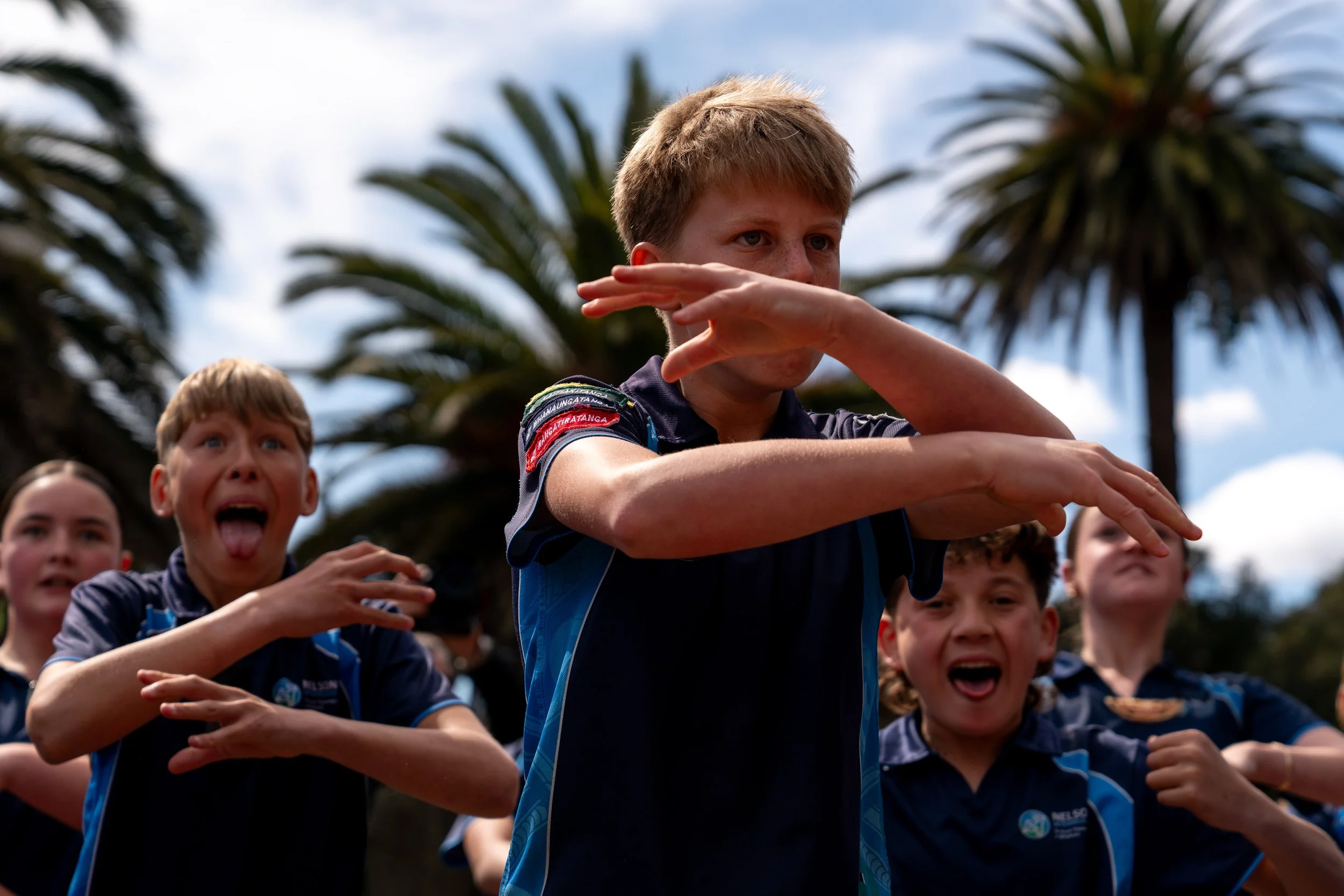 Group of children outdoors, wearing blue sports uniforms, doing a dance or drill, with palm trees and cloudy sky in the background.