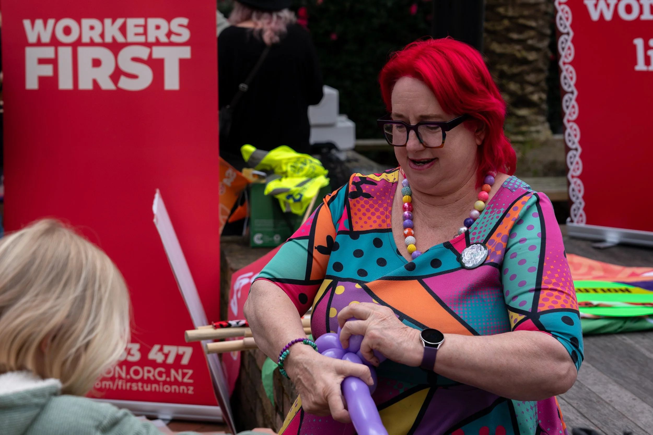 A woman with bright red hair, wearing colorful clothing and a multicolored necklace, inflates a purple balloon at an outdoor event with a red banner that reads 'WORKERS FIRST' in the background.