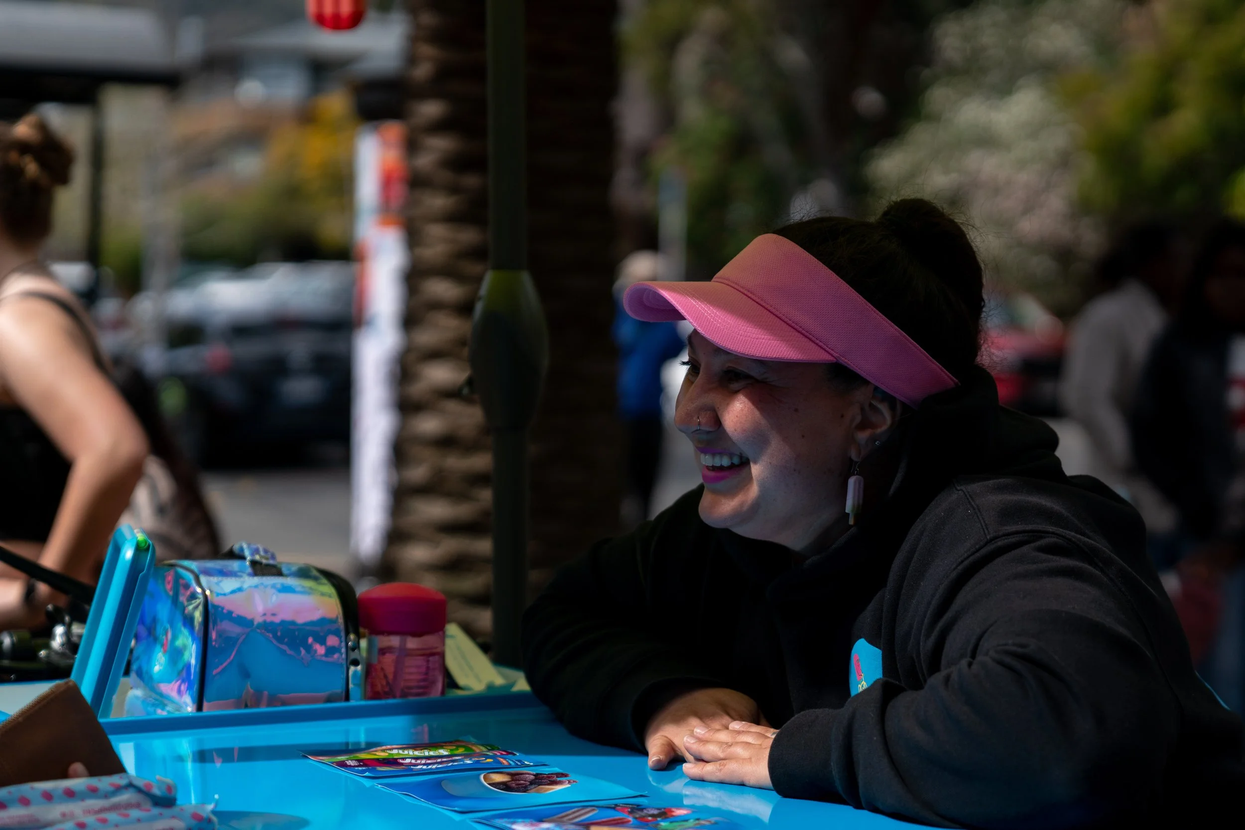 A woman in a pink visor smiling and talking at a table outdoors, with other people and trees in the background.