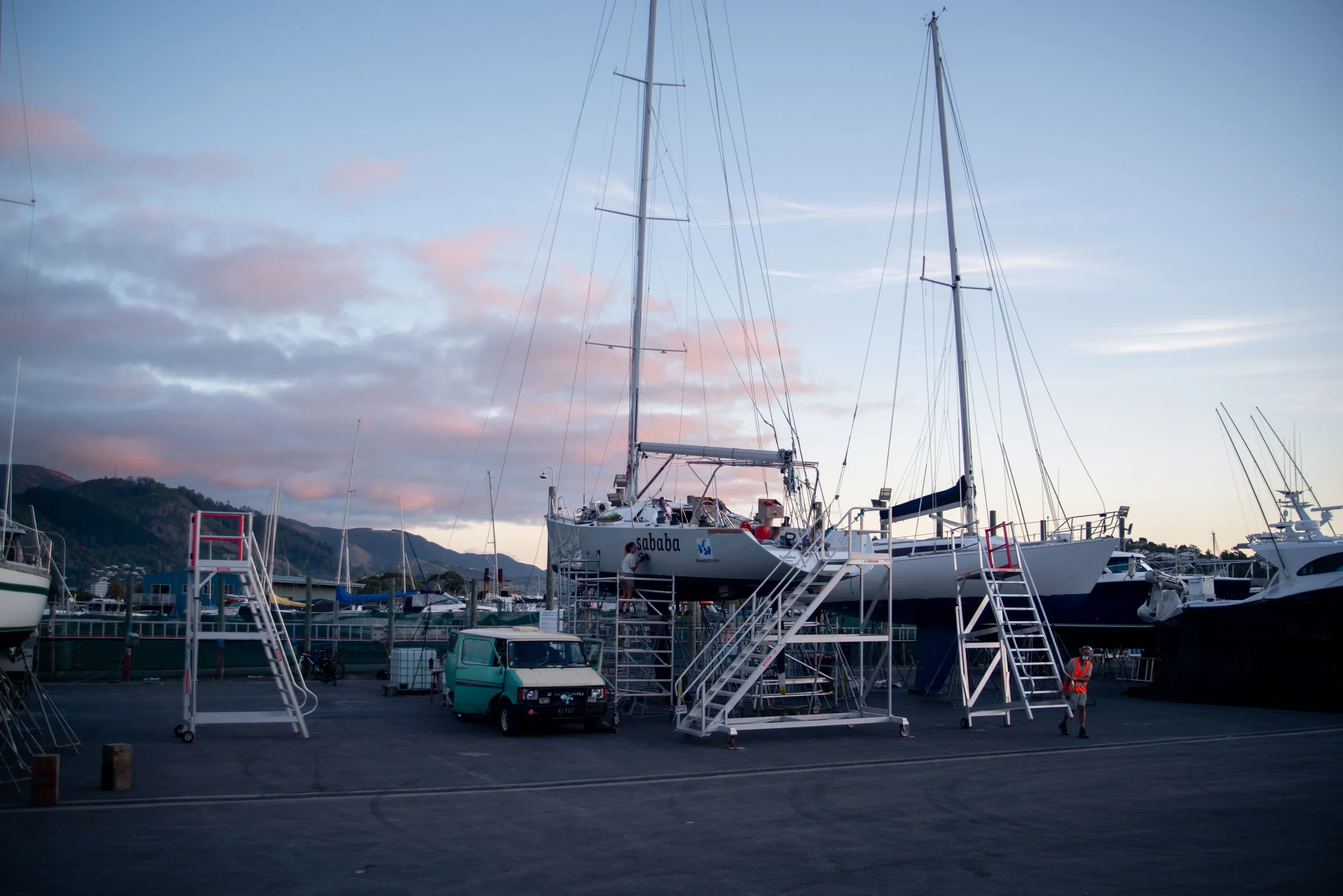 A marina during sunset with sailboats on dry dock, a person in a safety vest walking, and a small turquoise vehicle parked.