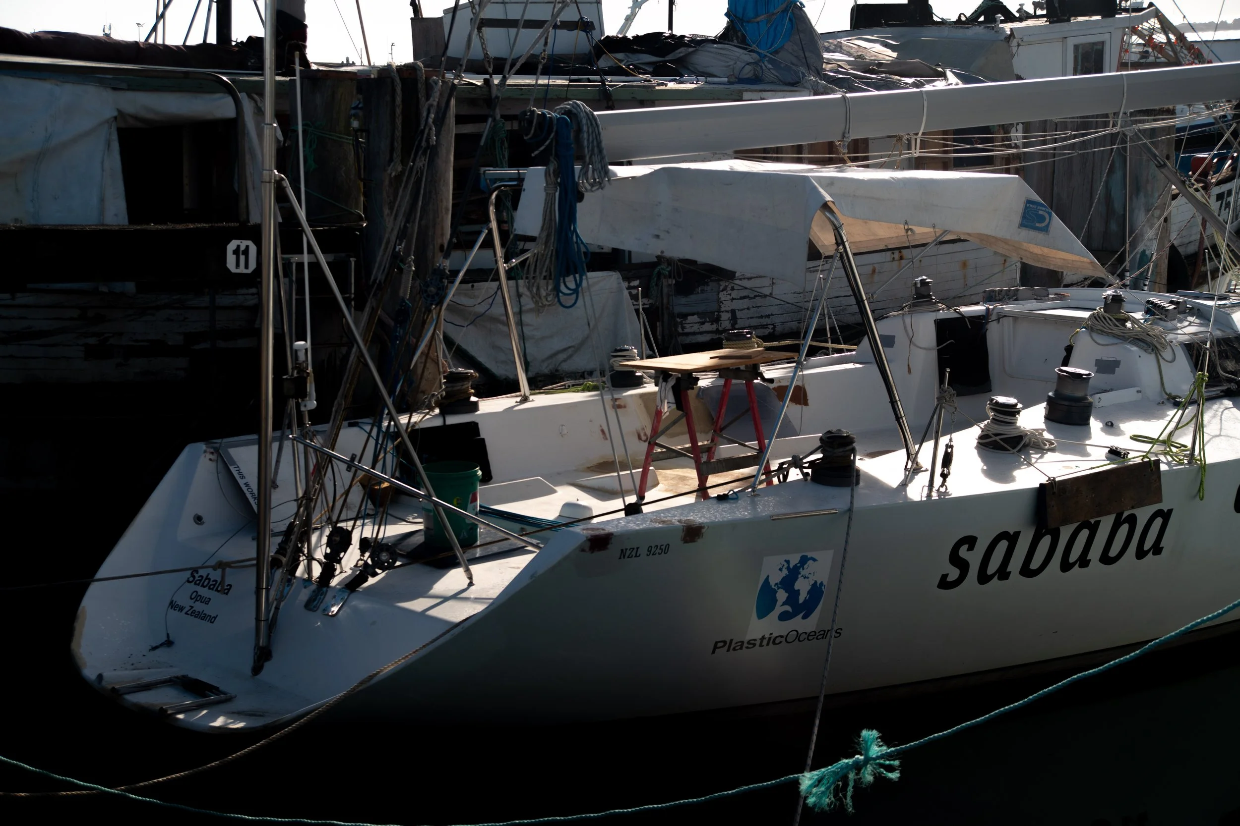 A sailboat named Sababa docked at a marina with other boats, showing ropes, winches, and a chart table, with a New Zealand registration and a Plastic Oceans sticker.