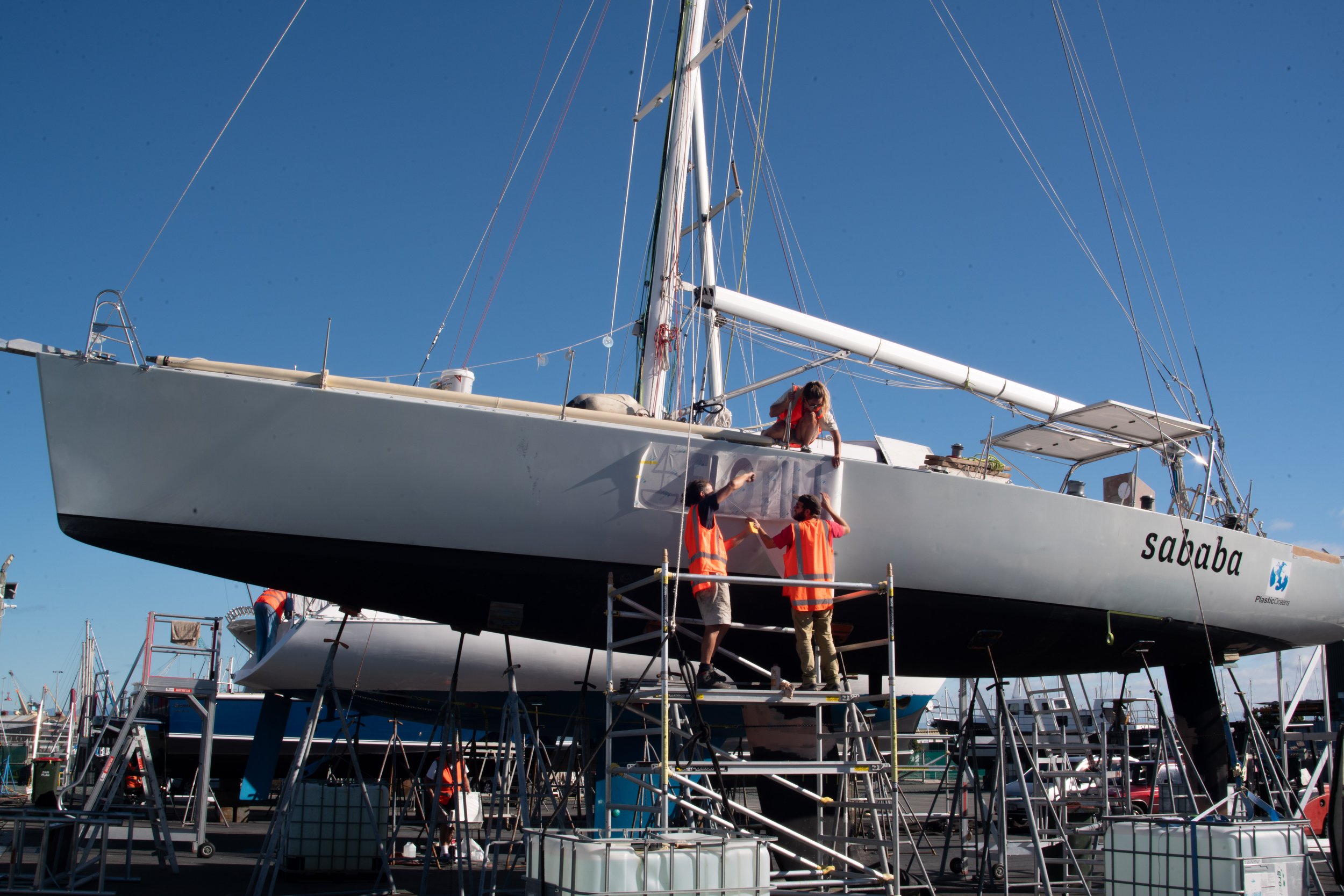 People in safety vests working on a large white sailing yacht named 'Sababa' in a boatyard, with scaffolding around the boat, under a clear blue sky.